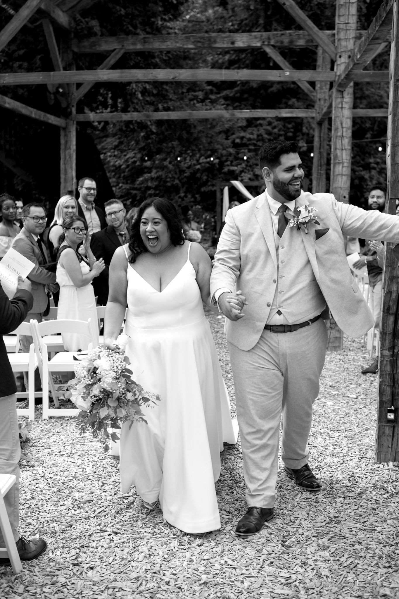 A joyful newlywed couple walks down the aisle at an outdoor rustic wedding ceremony while guests celebrate.