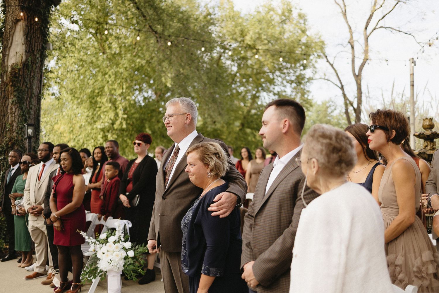 Wedding guests gather outdoors for a celebration under tree canopy.