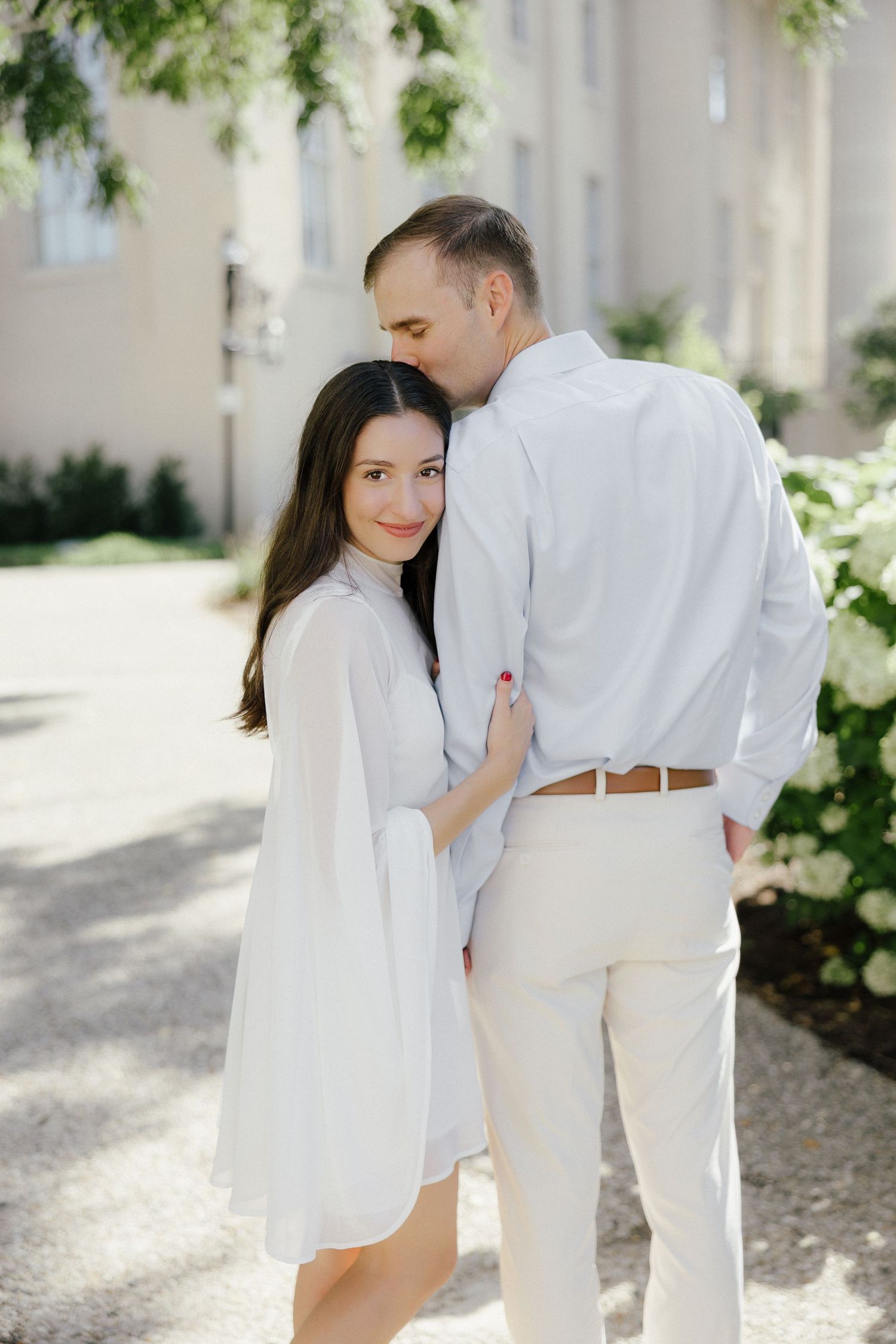 Woman in white dress leaning into man in light shirt, standing near blooming hydrangeas outdoors.