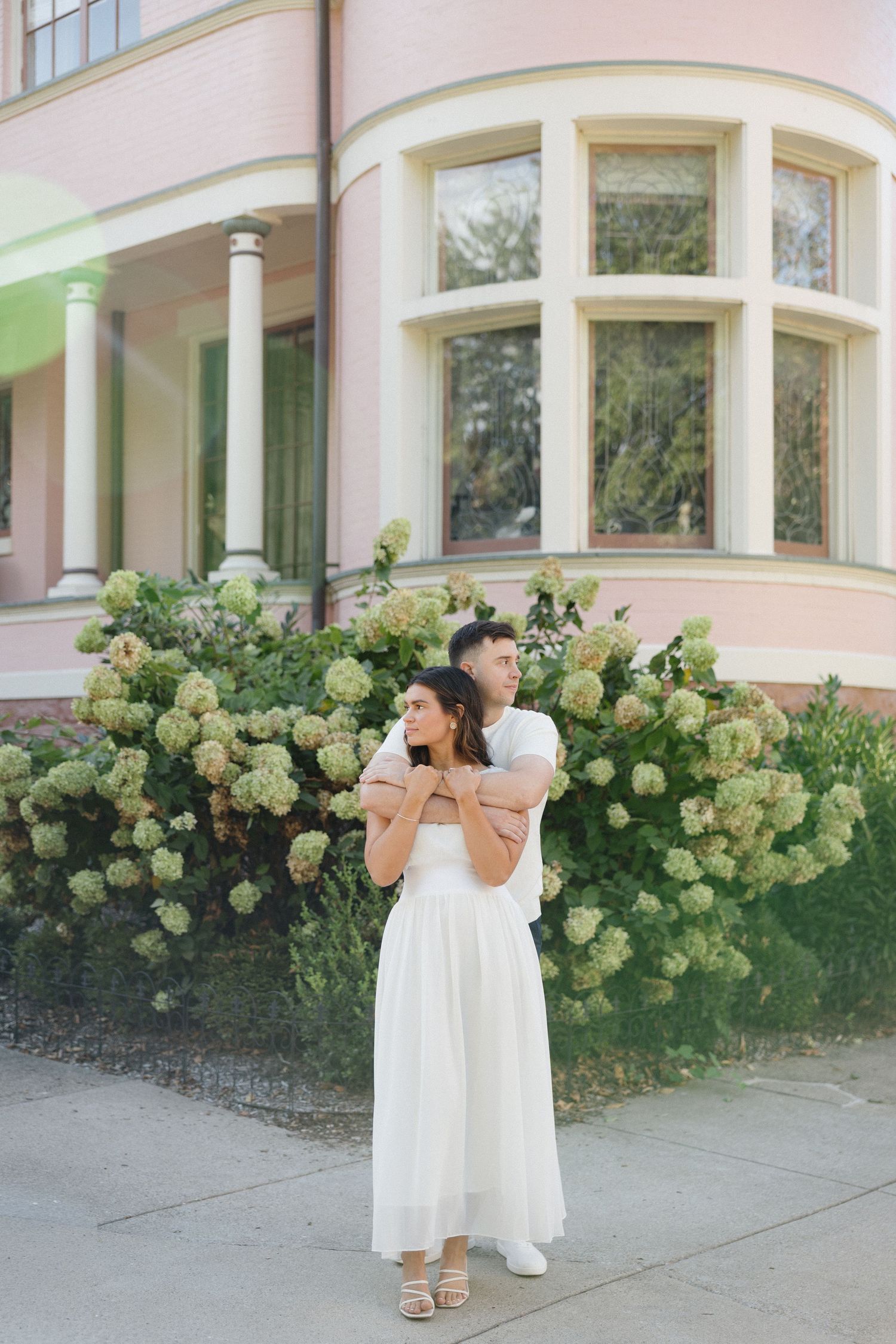 Couple embracing in front of blooming hydrangea bushes near a pink building, woman in white dress.