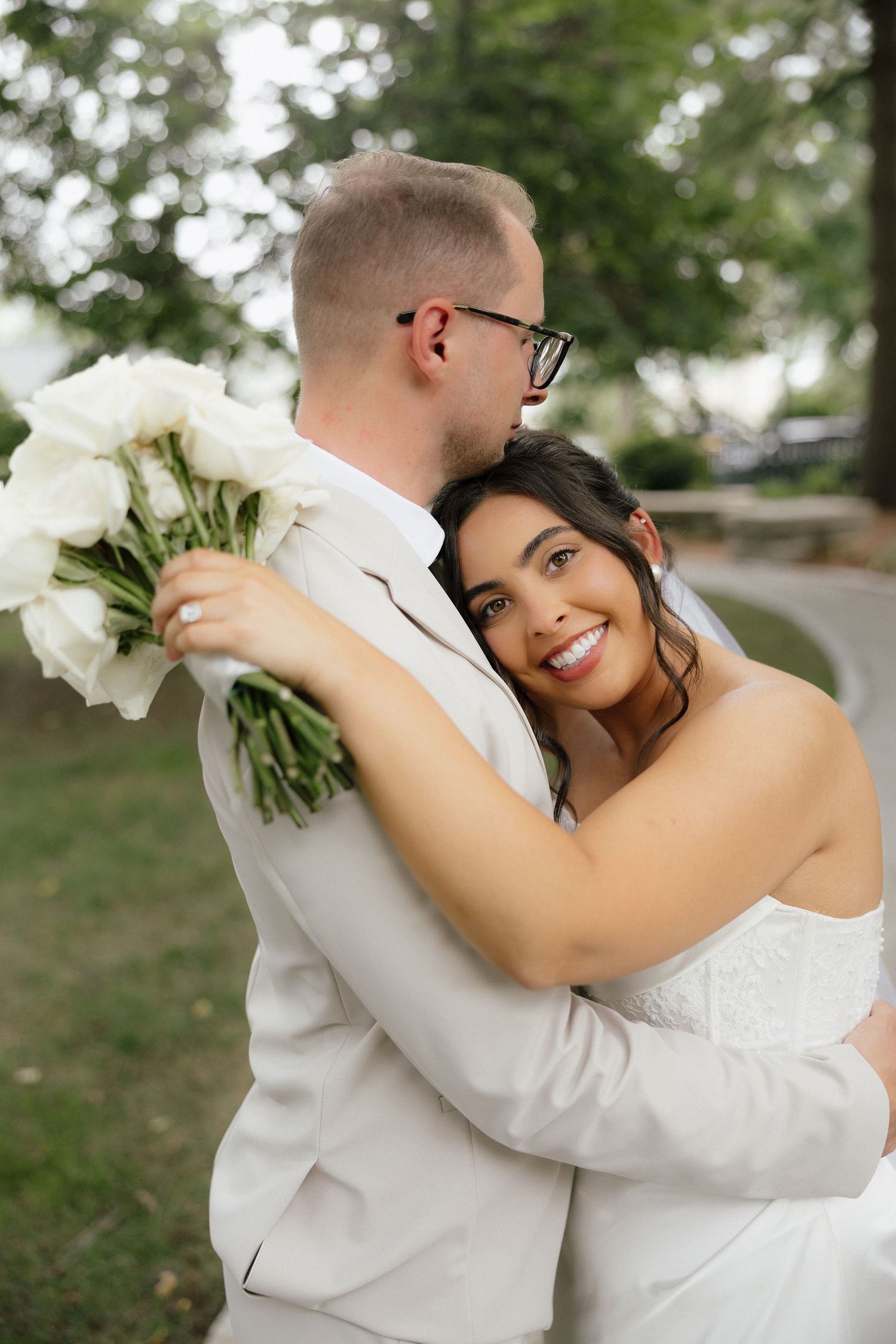 Happy bride in strapless gown embraces groom holding white floral bouquet in outdoor wedding portrait.