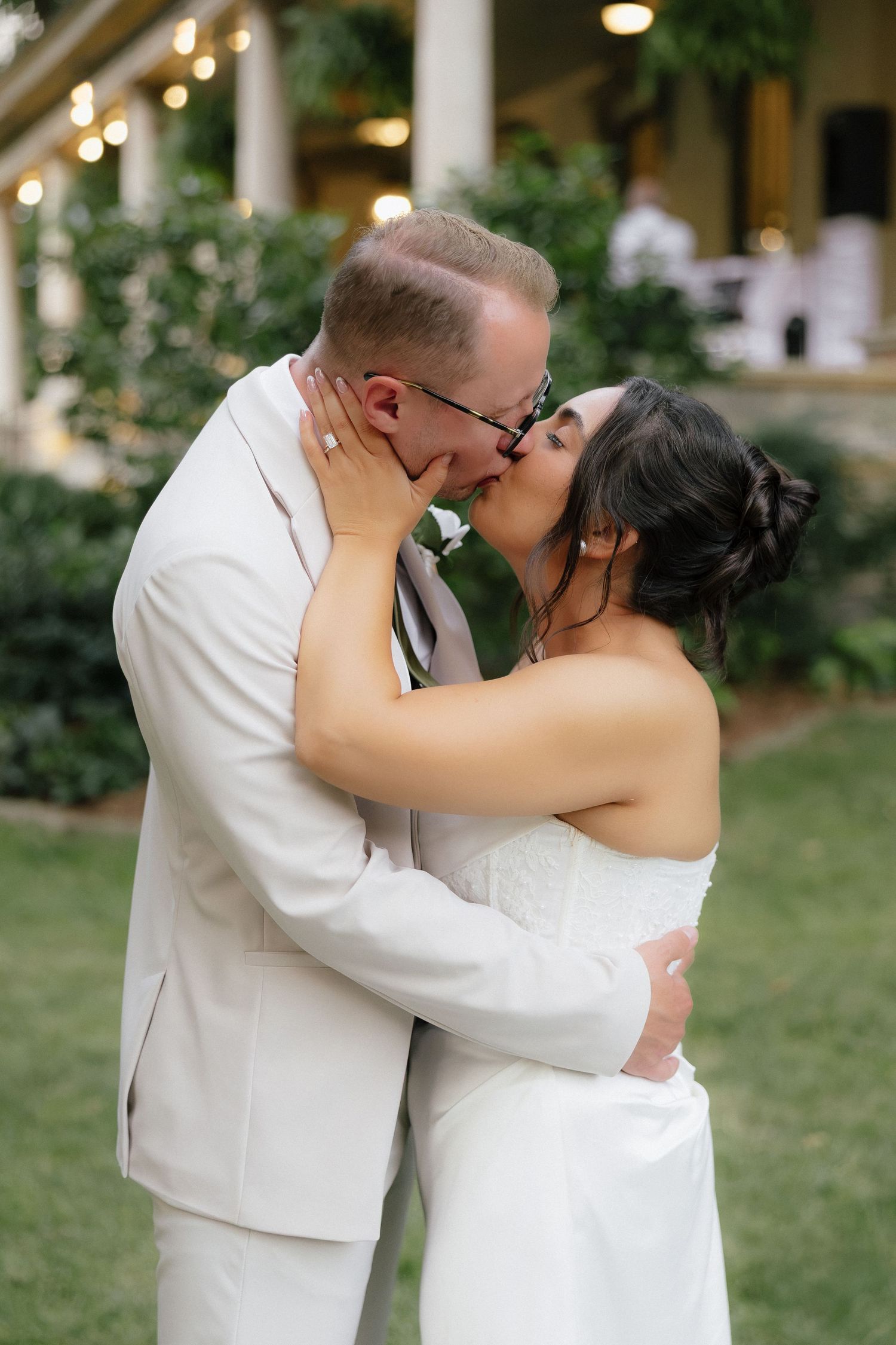 Bride and groom share passionate kiss outdoors with elegant venue columns and greenery in background.