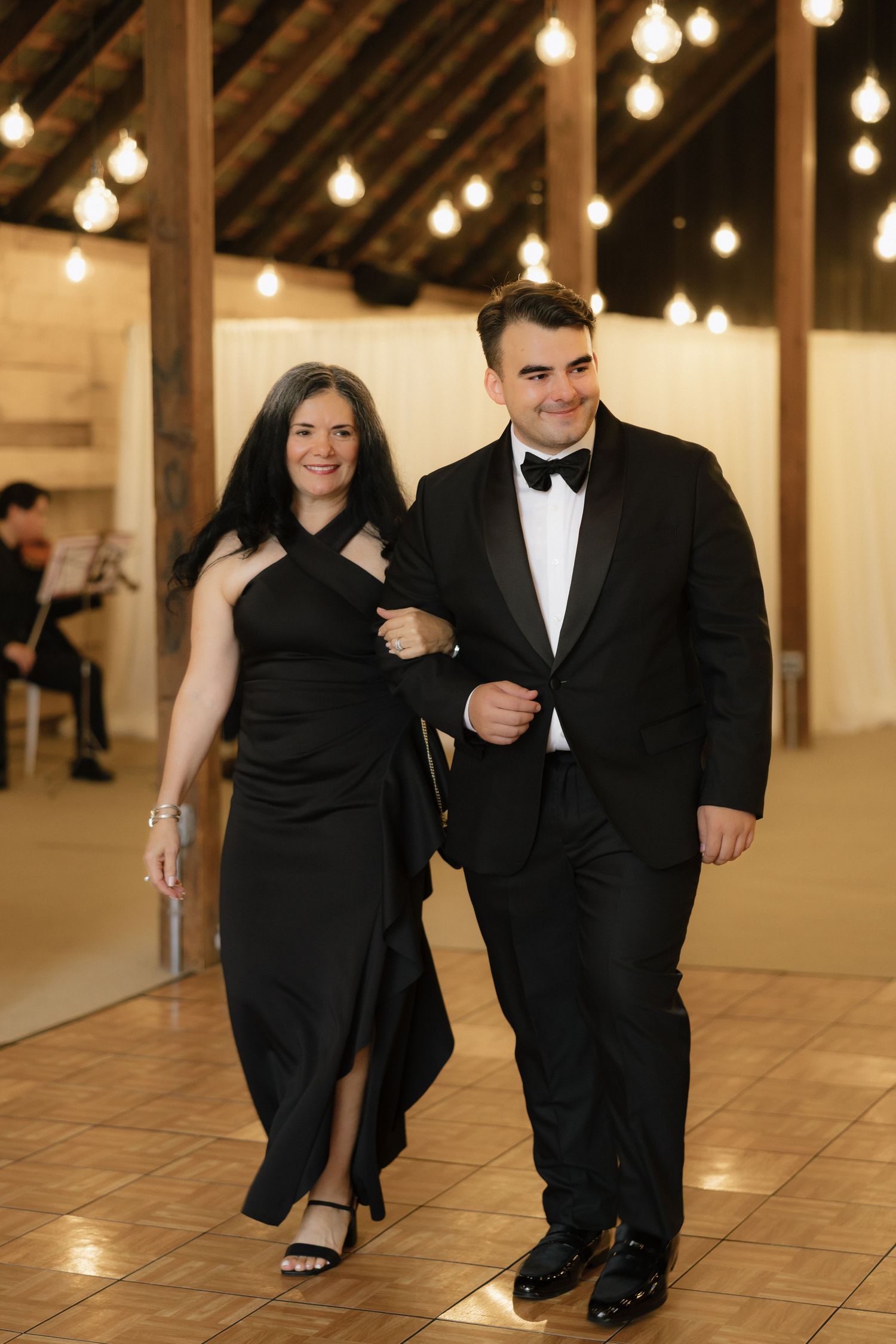 Groom in black tuxedo escorts woman in black gown across dance floor at wedding reception.