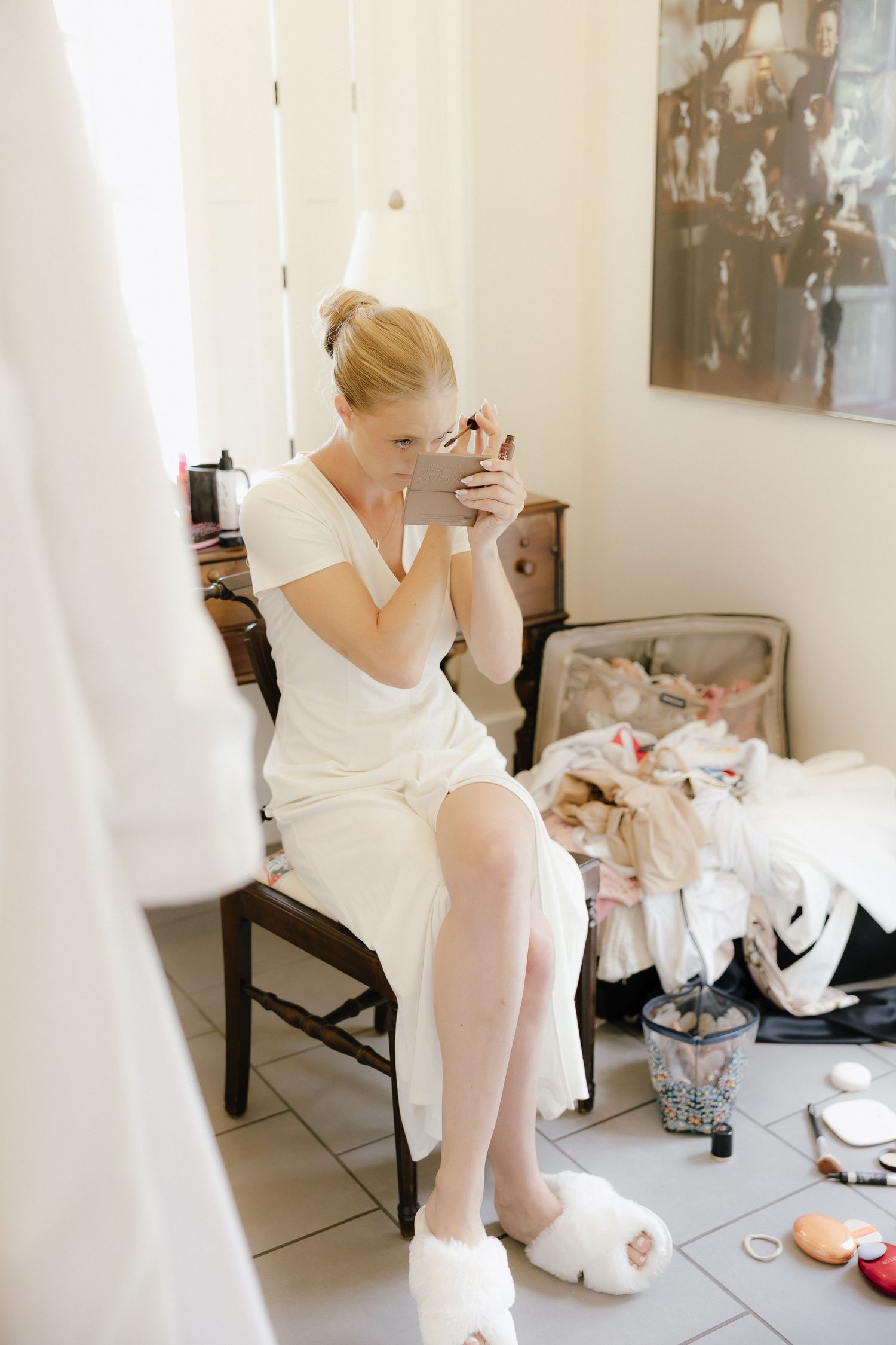 Bride in white dress applying makeup with a hand mirror, sitting in a chair surrounded by wedding preparations.