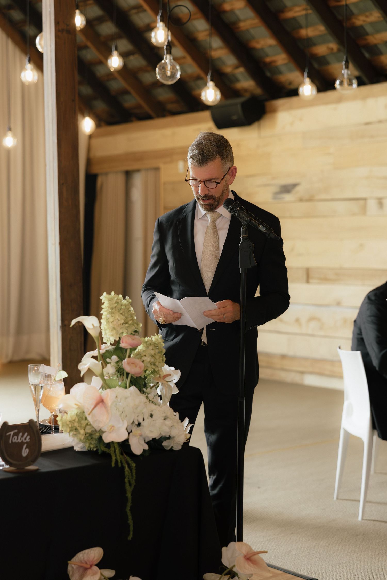 Man in dark suit delivering speech at microphone beside floral centerpiece at wedding reception.