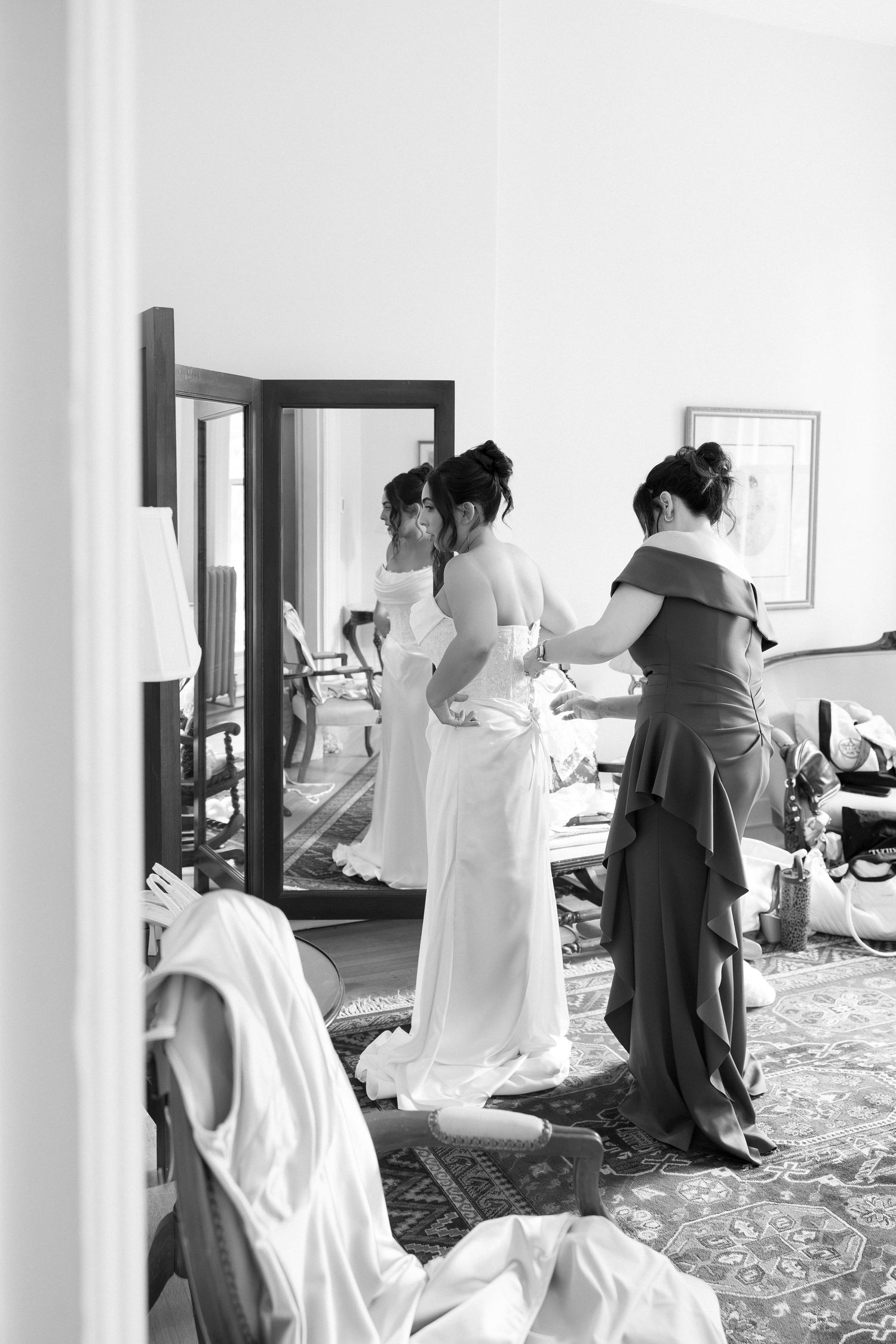 Black and white photo of a bride being helped into her wedding dress by a bridesmaid in front of a mirror.