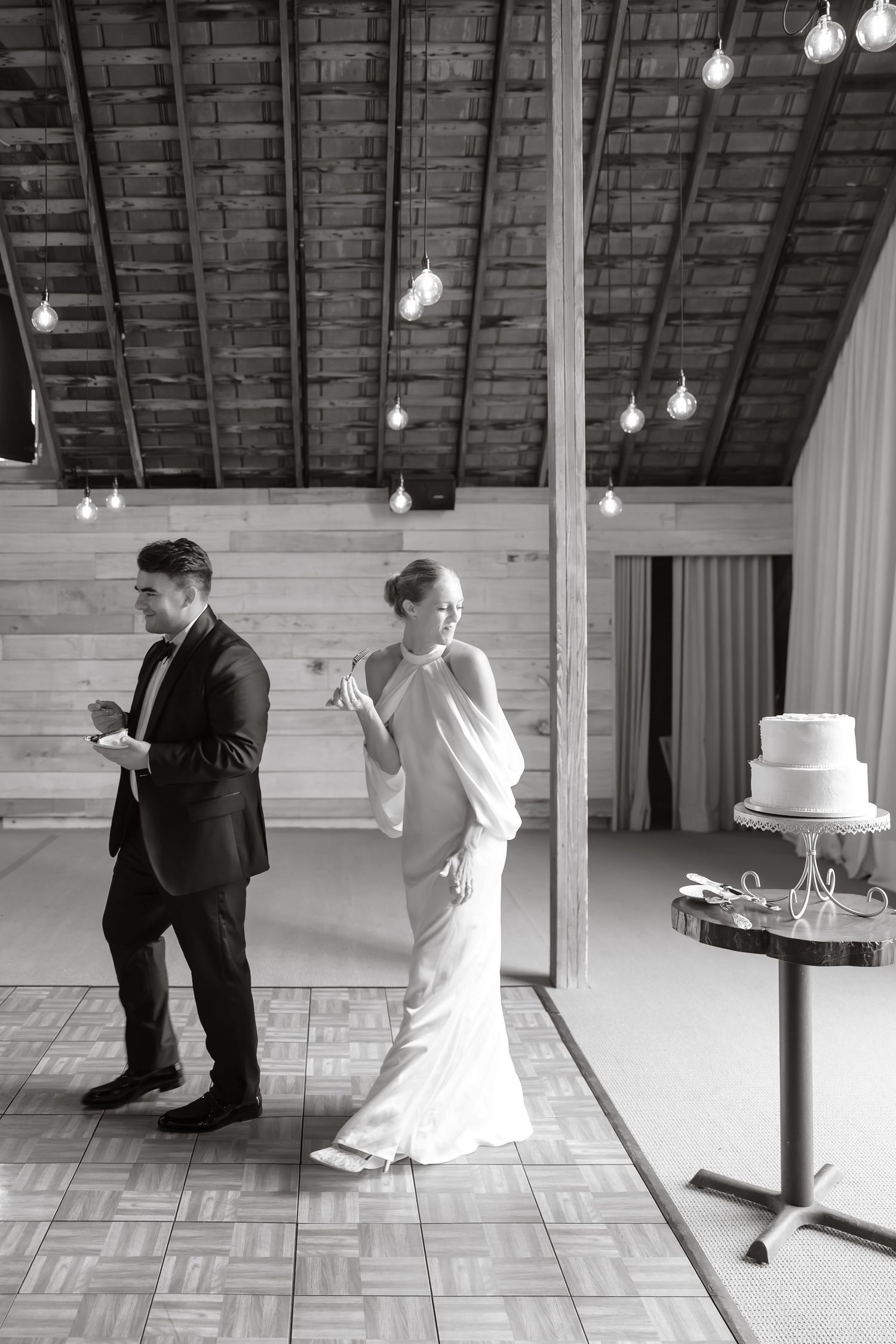 Black and white photo of bride and groom cutting wedding cake in rustic barn reception venue.