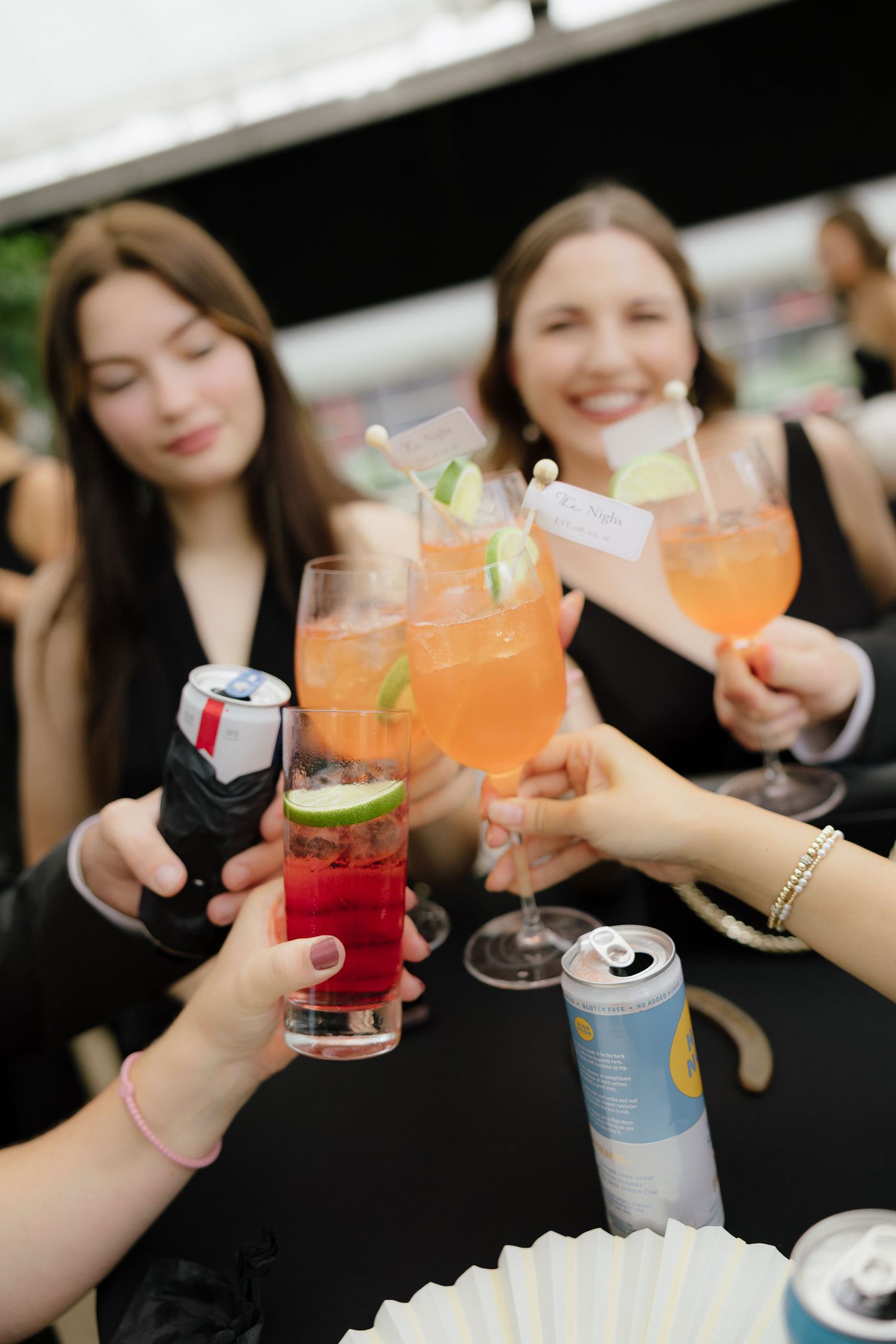 Group of women in black dresses toasting with colorful cocktails and canned drinks at an outdoor event.