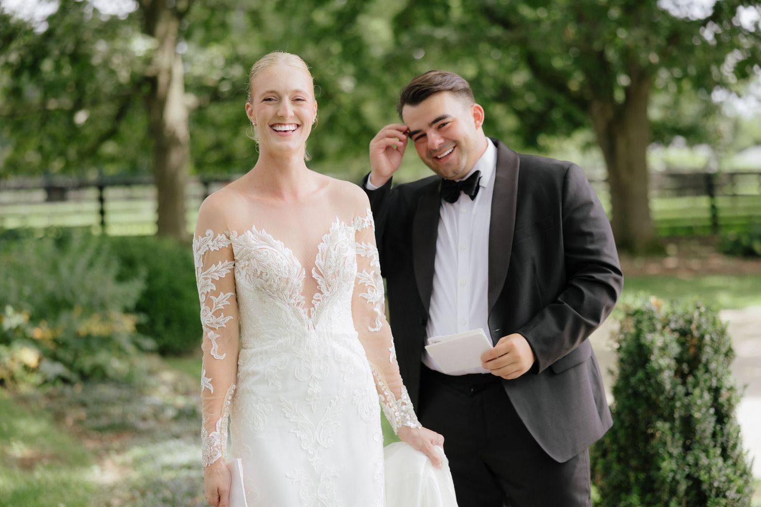 Bride in lace long-sleeve wedding gown stands with groom in black tuxedo outdoors at elegant garden wedding venue.