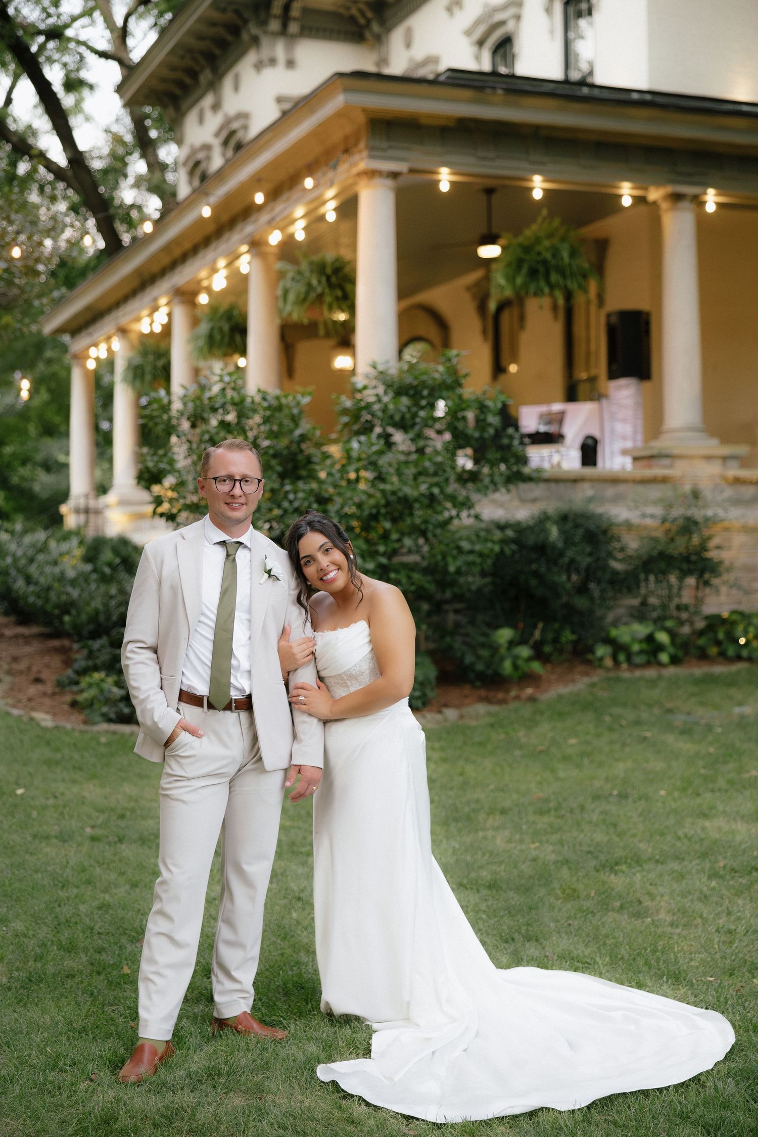 Bride and groom posing together on green lawn in front of elegant venue with string lights and columns.