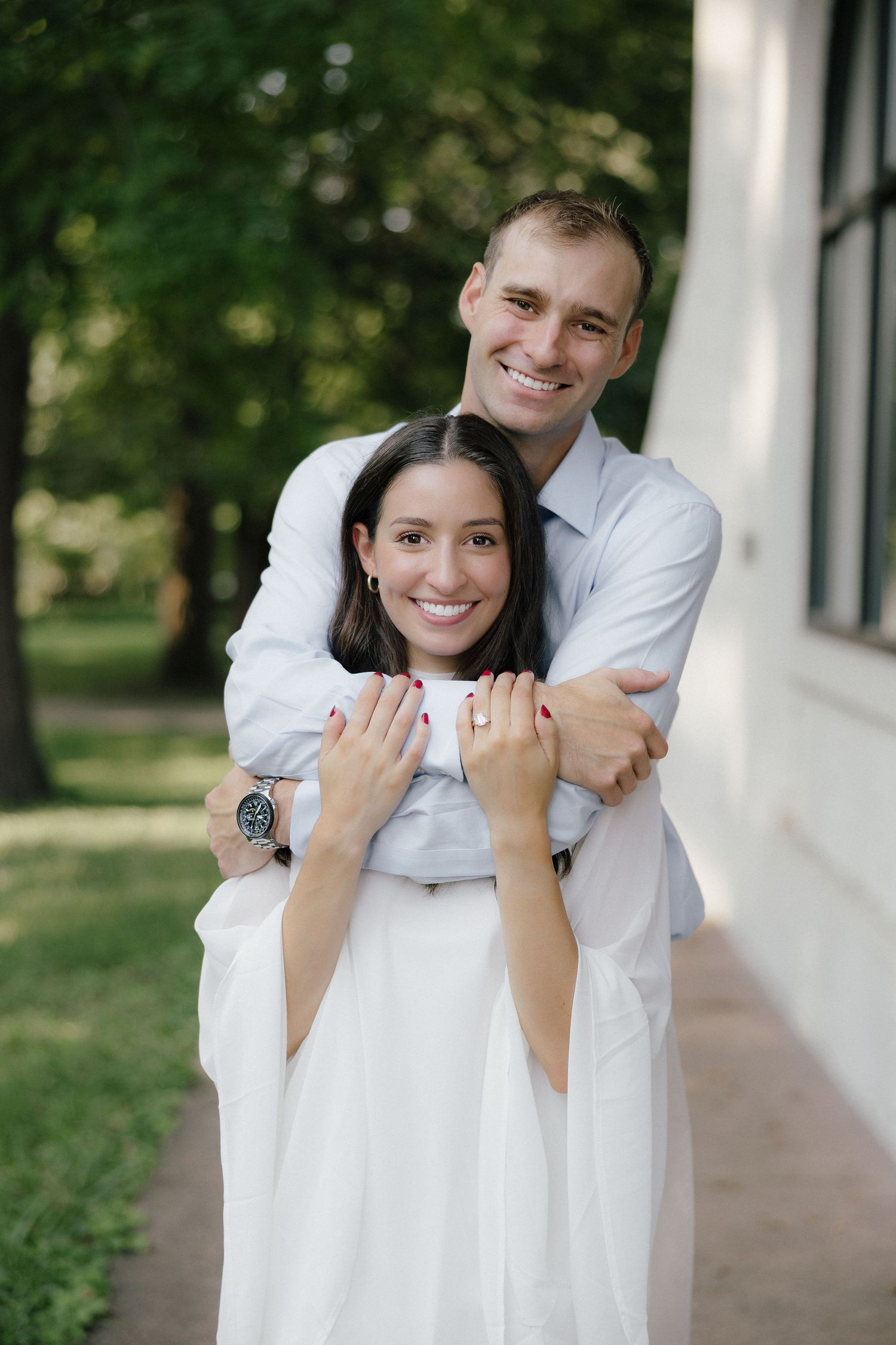 Happy couple embracing outdoors, man hugging woman from behind as she displays engagement ring, surrounded by green trees.