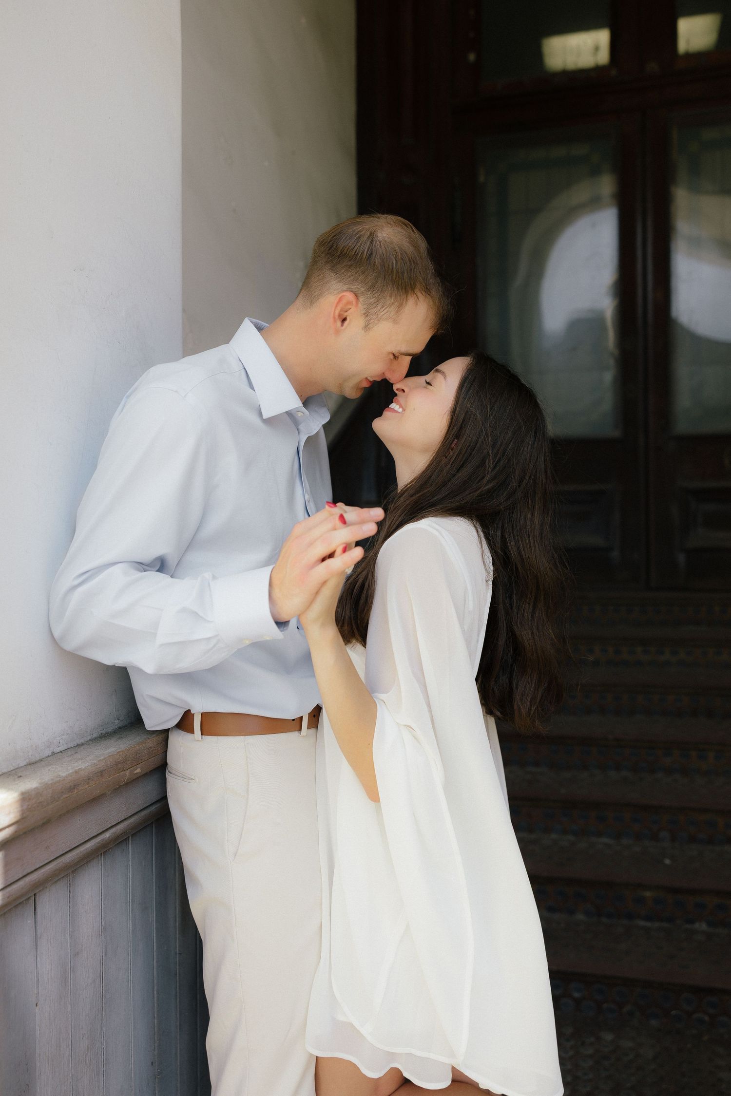 Romantic couple leaning in for a kiss while holding hands against a classic stone building column.