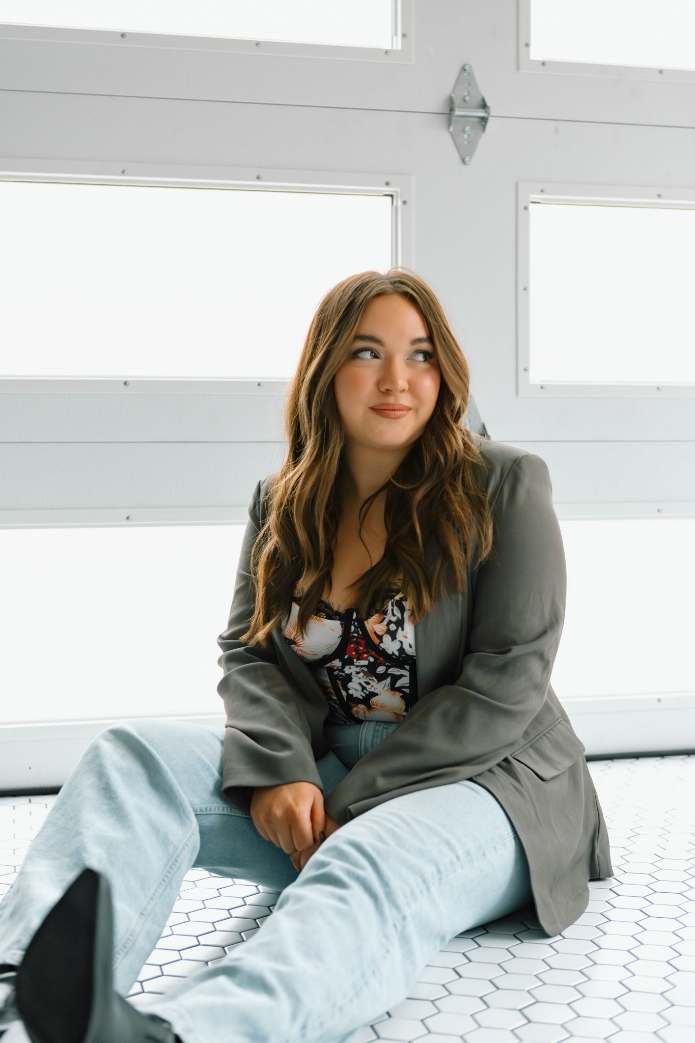 Young woman in gray blazer and light jeans sits on tile floor, gazing thoughtfully to the side near bright windows.