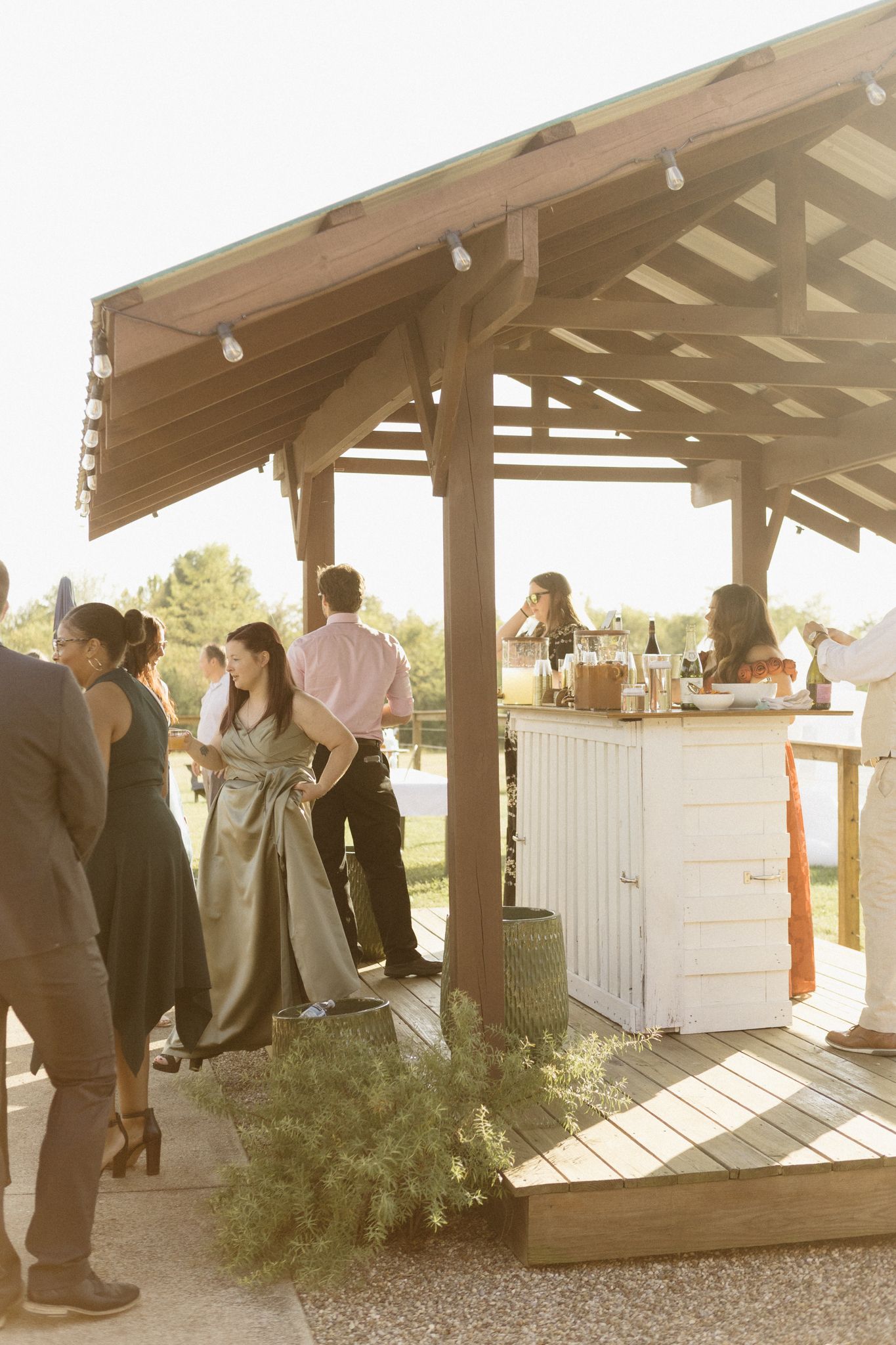 People gather under a wooden pavilion at an outdoor event venue with white bar counters.