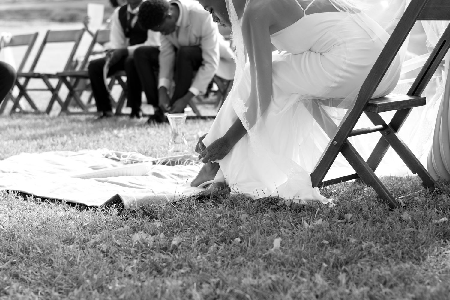 Wedding guests seated in folding chairs casting shadows on grass in black and white.