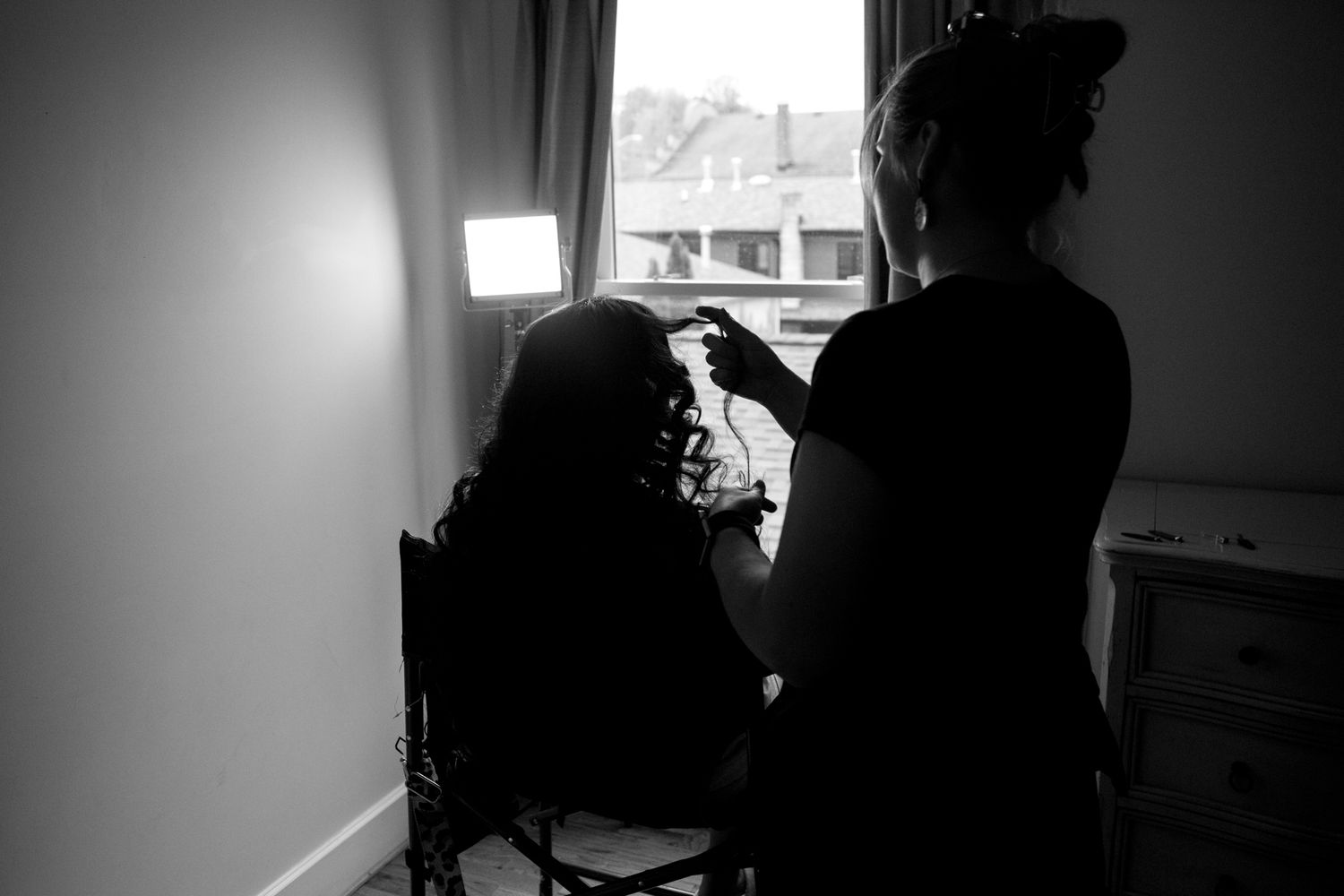 A silhouette stands by a window in a dark room working at an antique sewing machine.