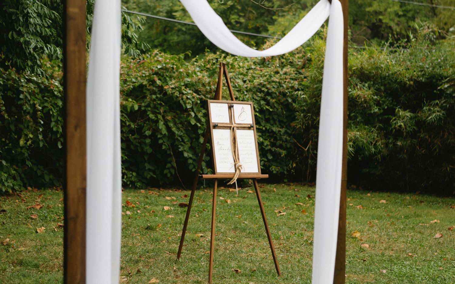 A wooden easel with a white seating chart stands beneath elegant white fabric draping at an outdoor wedding venue.