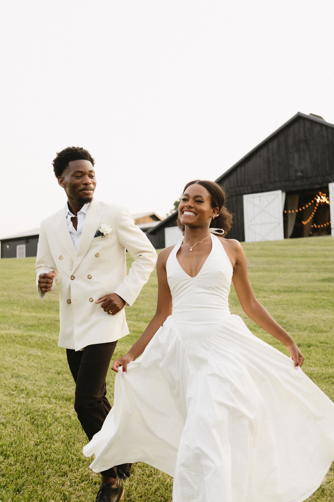 Joyful newlyweds dance and twirl in elegant white attire on a grassy lawn with a barn in the background.