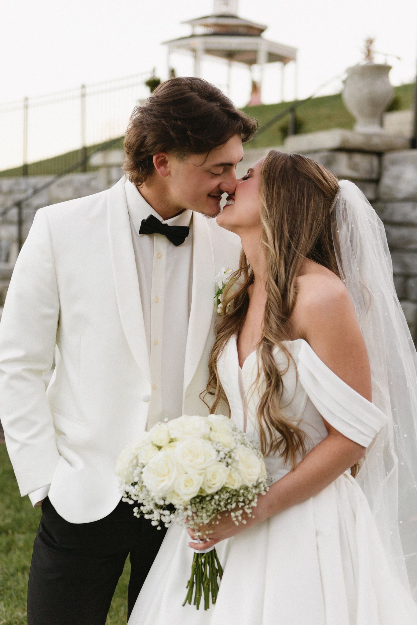 A romantic wedding couple sharing a kiss outdoors in formal attire with a white bouquet.