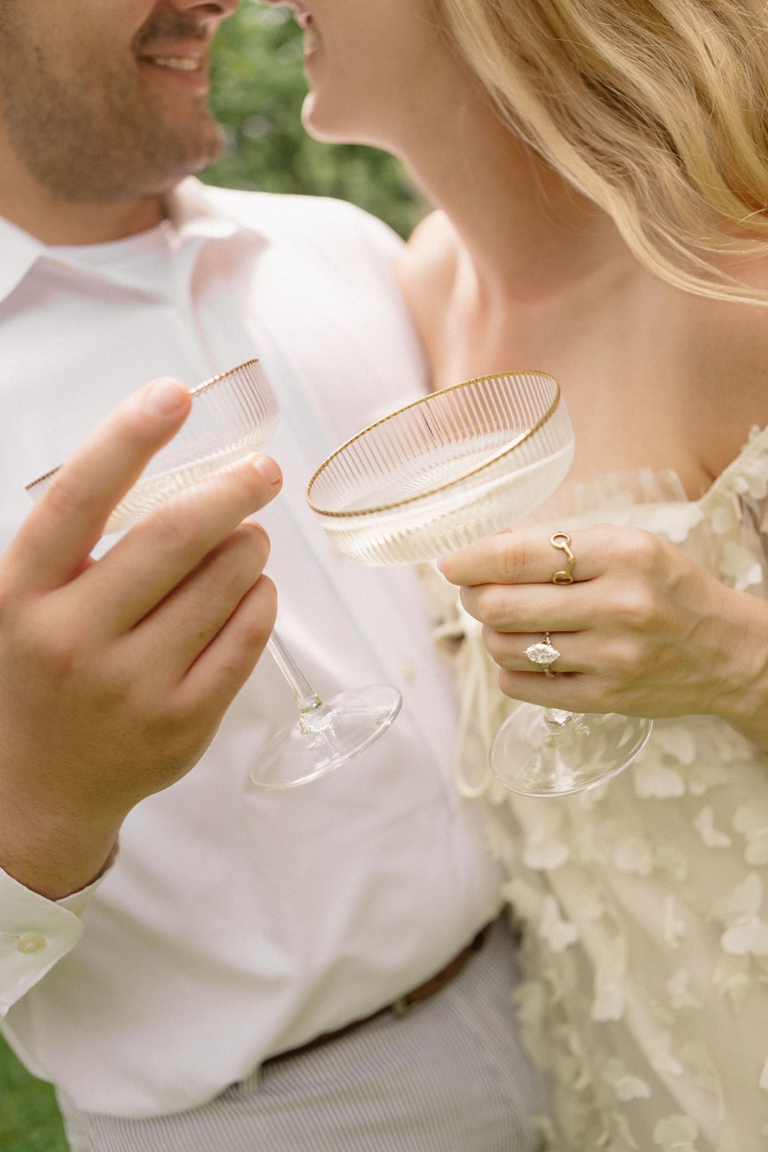 A couple in wedding attire clinks elegant champagne coupe glasses outdoors, with a diamond engagement ring visible on the bride's hand.