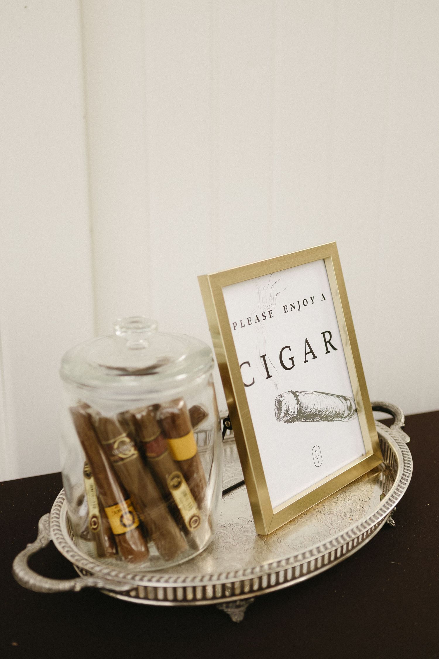 Elegant display of cigars in a glass jar with a framed sign reading 'Please Enjoy a Cigar' on a silver tray.