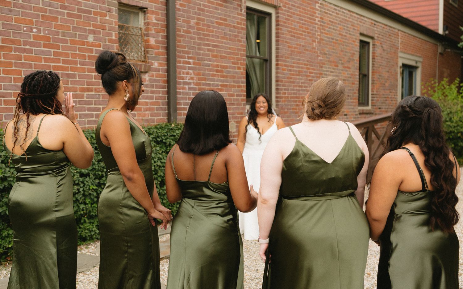 Wedding party wearing coordinated olive green dresses stand together in front of a brick building exterior.