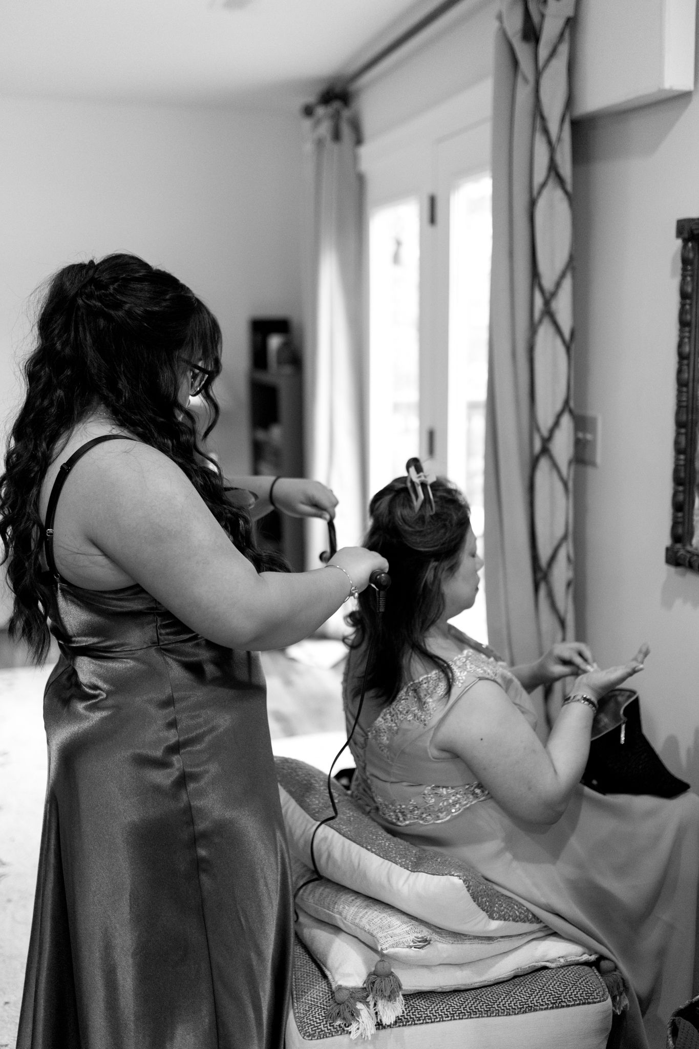 A bridesmaid in a dark dress helps style another person's hair during wedding preparations in a sunlit room.