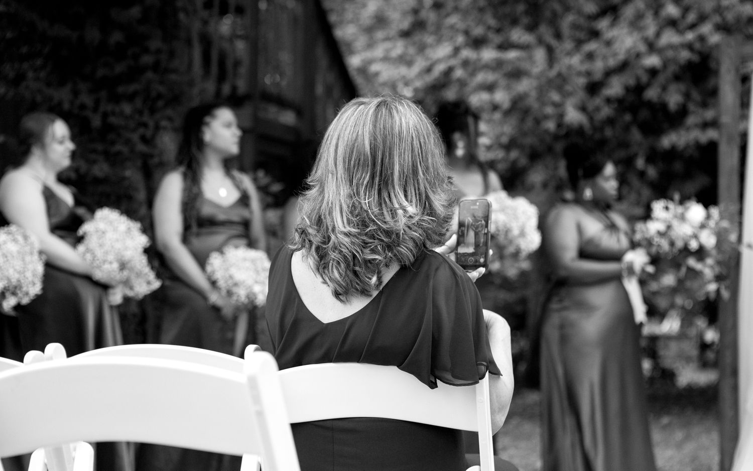 Black and white photo of a wedding ceremony with seated guests viewing bridesmaids holding bouquets in formal dresses.