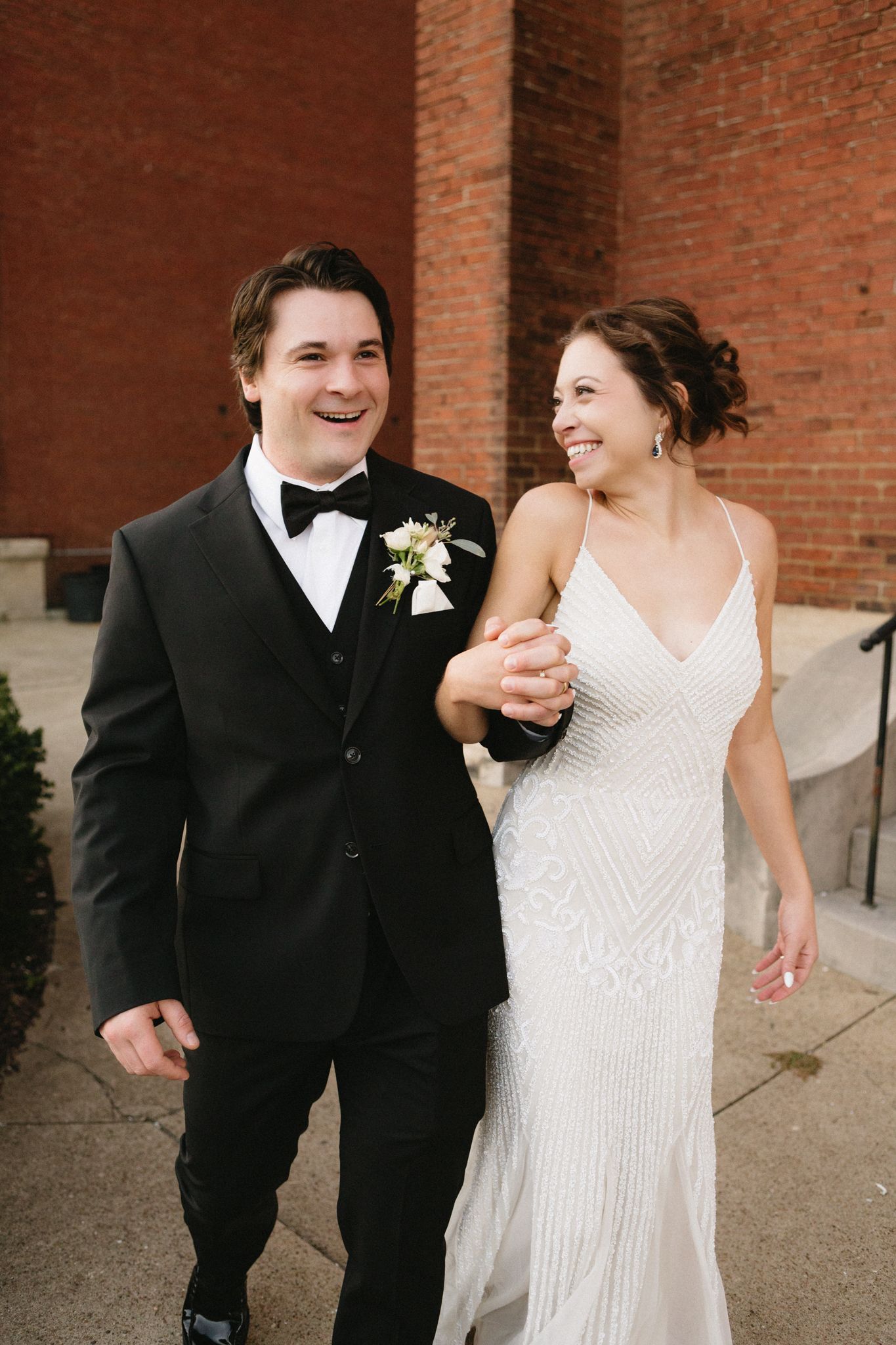 A couple in wedding attire laughs while walking together against a brick wall.