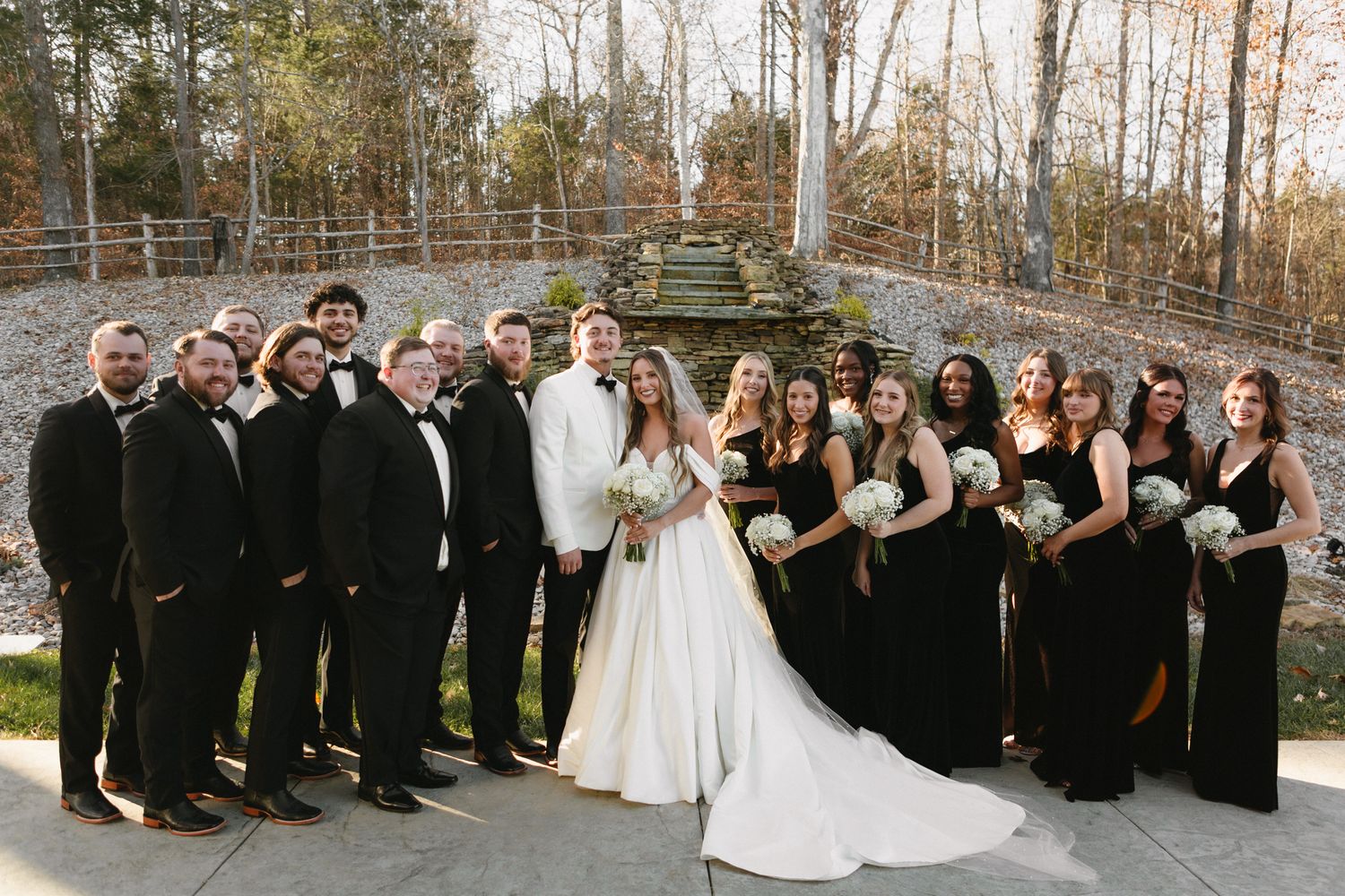Wedding party poses outdoors in formal black and white attire against a stone wall backdrop in autumn.