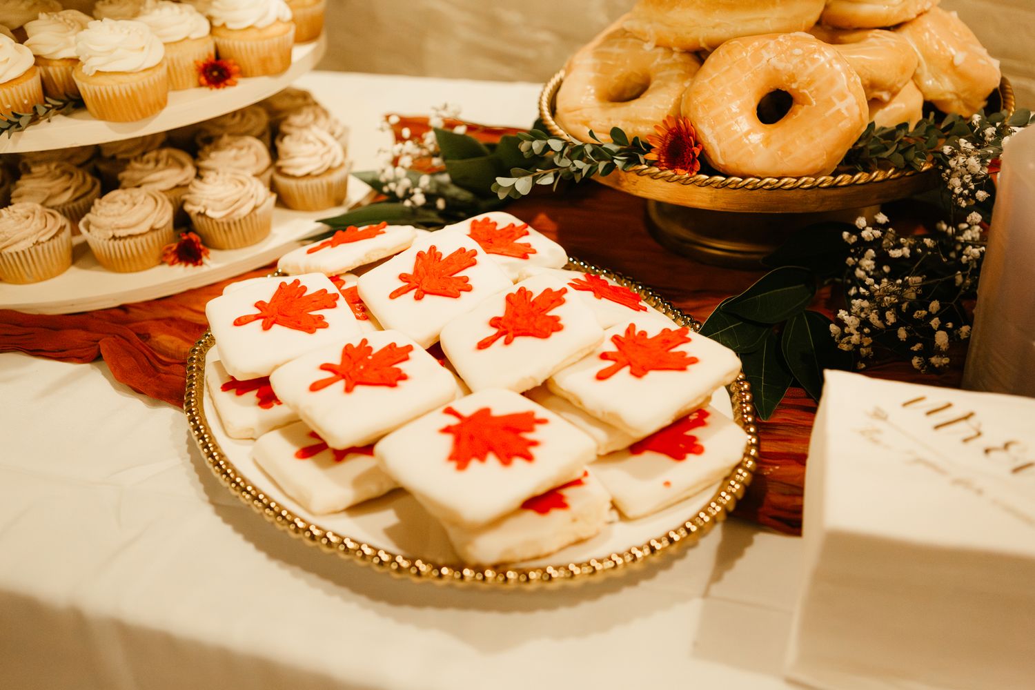 Reception dessert table with decorated cookies featuring red maple leaves and plated donuts on elegant stands.