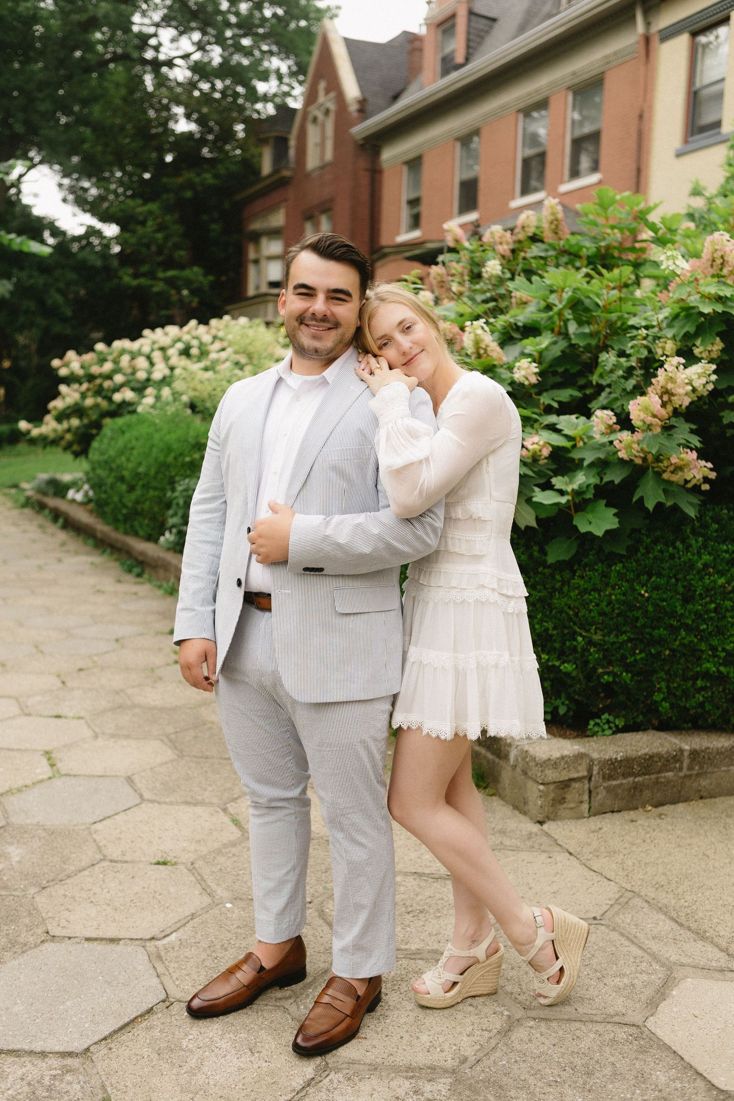 Couple posing outdoors in elegant attire, man in striped suit and woman in white dress, with lush garden background.