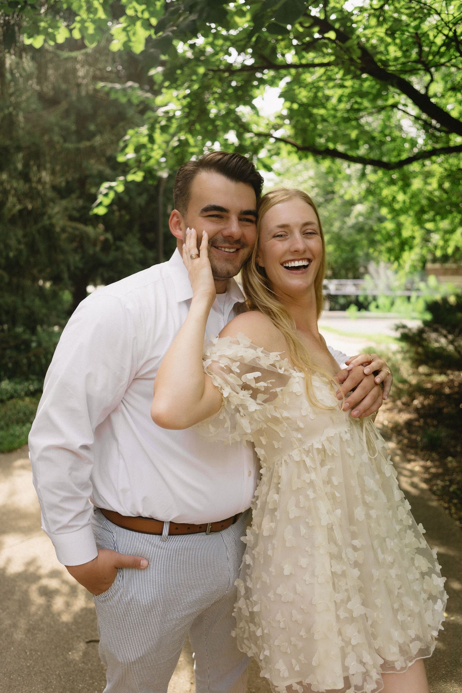 Joyful couple laughing outdoors in white outfits under green trees on a sunny day.
