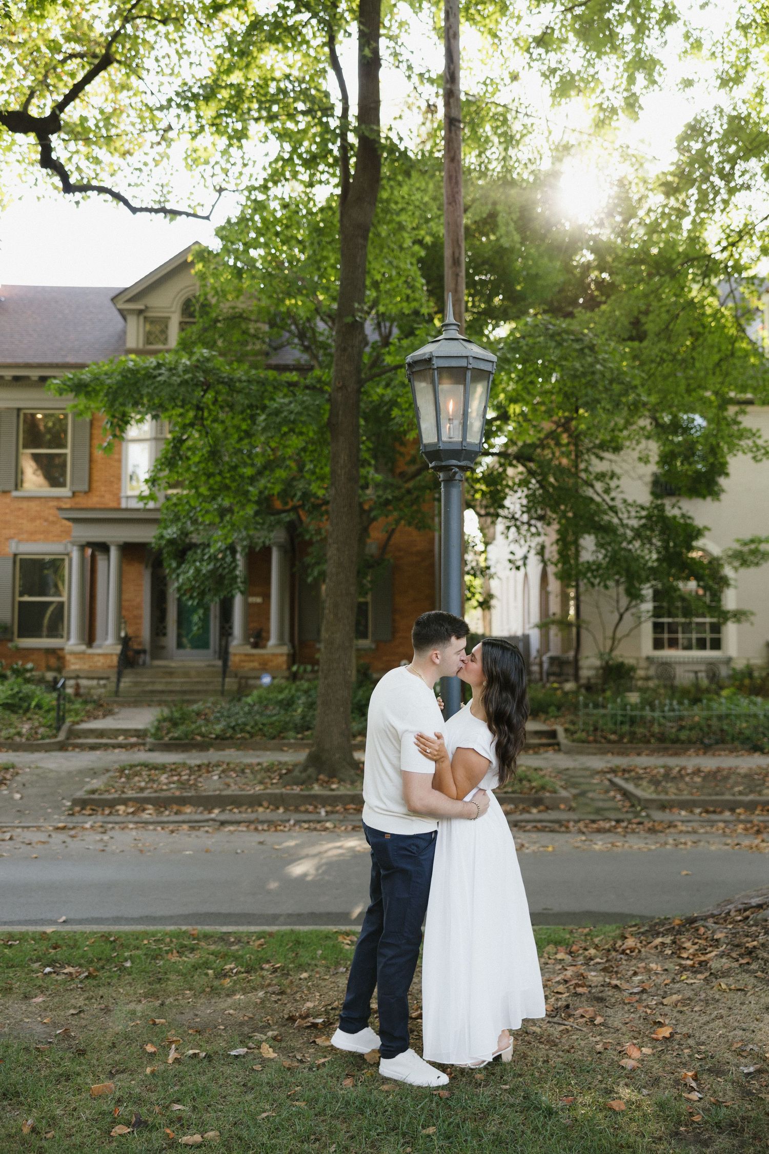 Couple sharing a kiss near a vintage street lamp with sunlit trees and historic homes in the background.
