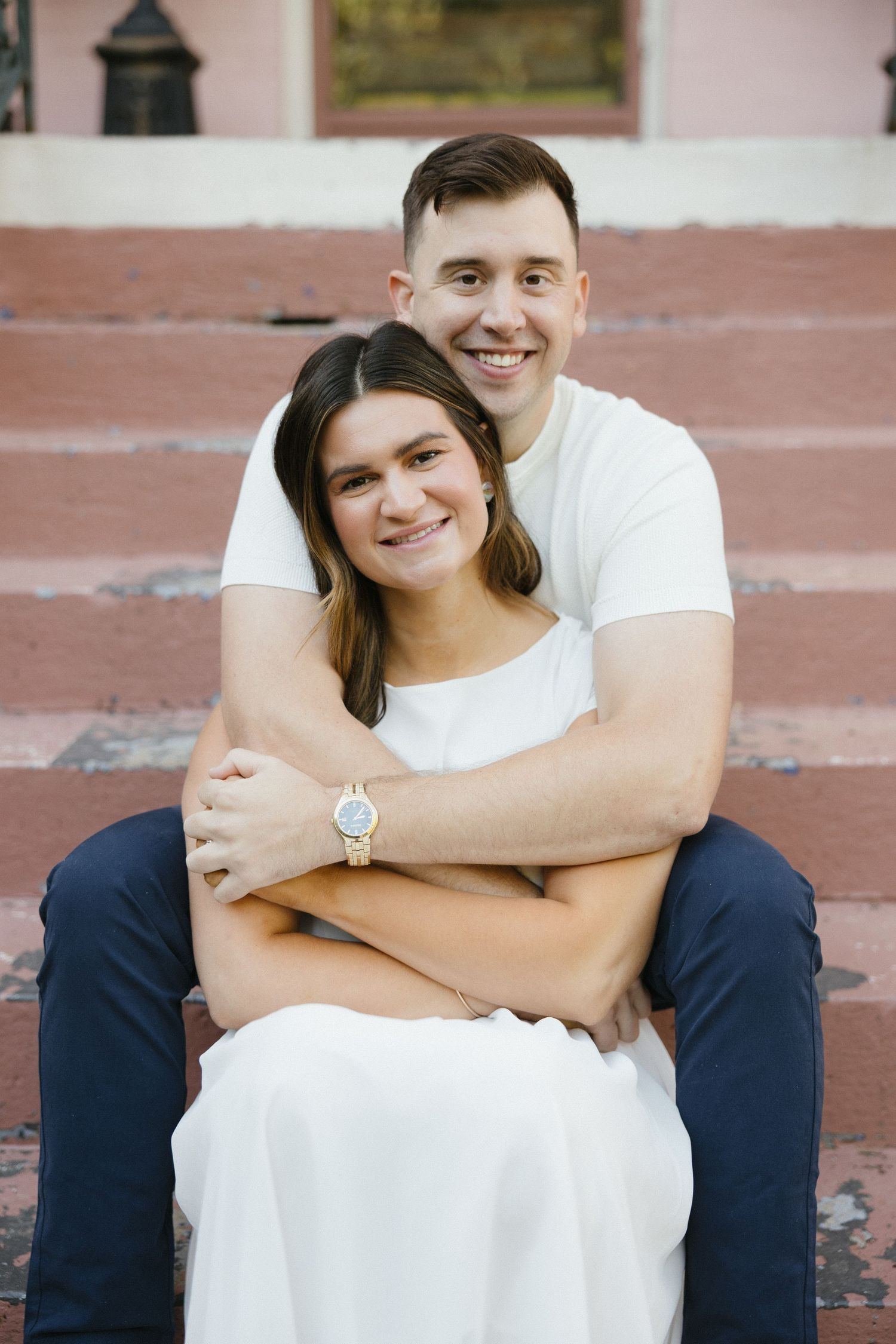 Smiling couple sitting on pink steps, man embracing woman from behind in a warm outdoor portrait.