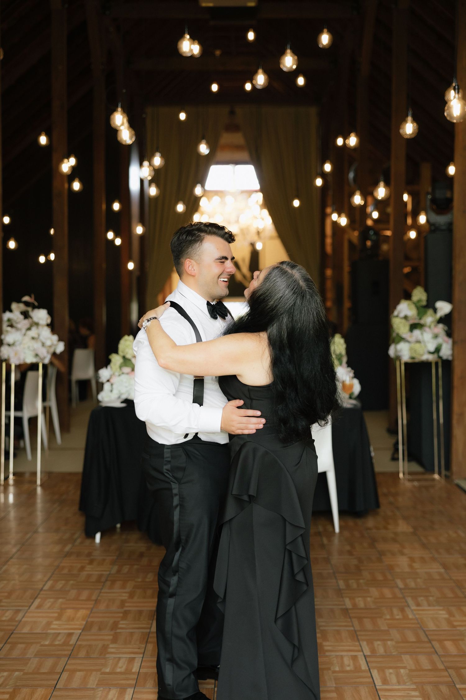 Groom dances with dark-haired woman in black dress at warmly lit barn wedding reception.