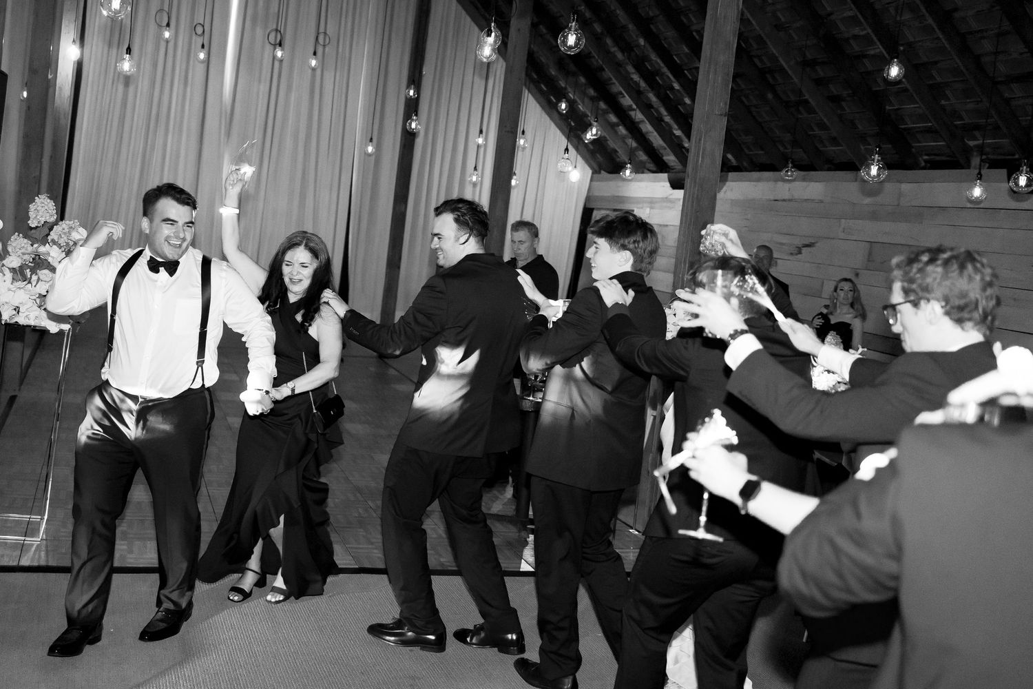 Black and white photo of wedding guests dancing energetically in barn reception venue with Edison lights.