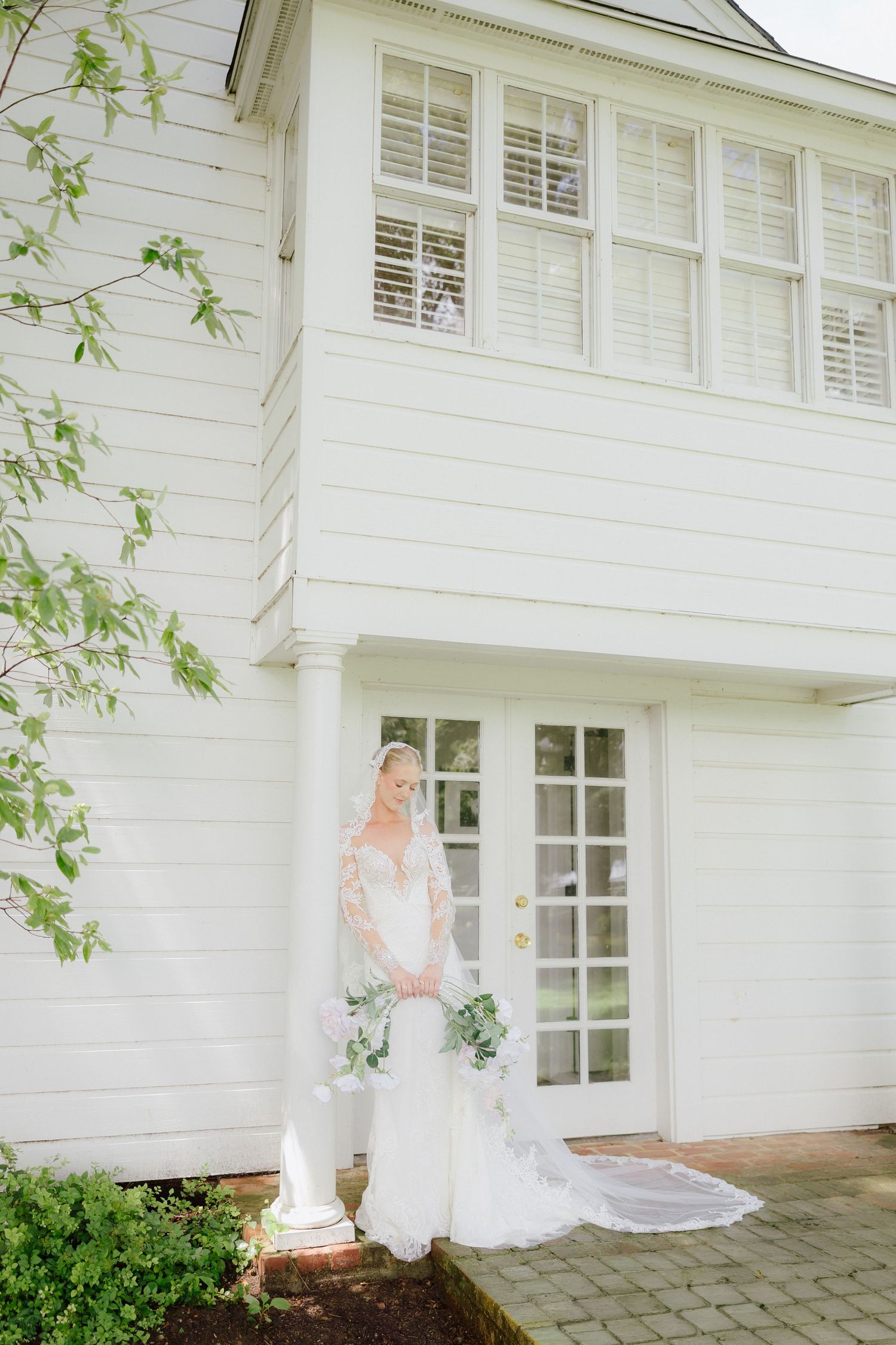 Bride in lace wedding gown and veil holding bouquet outside white colonial building with French doors.