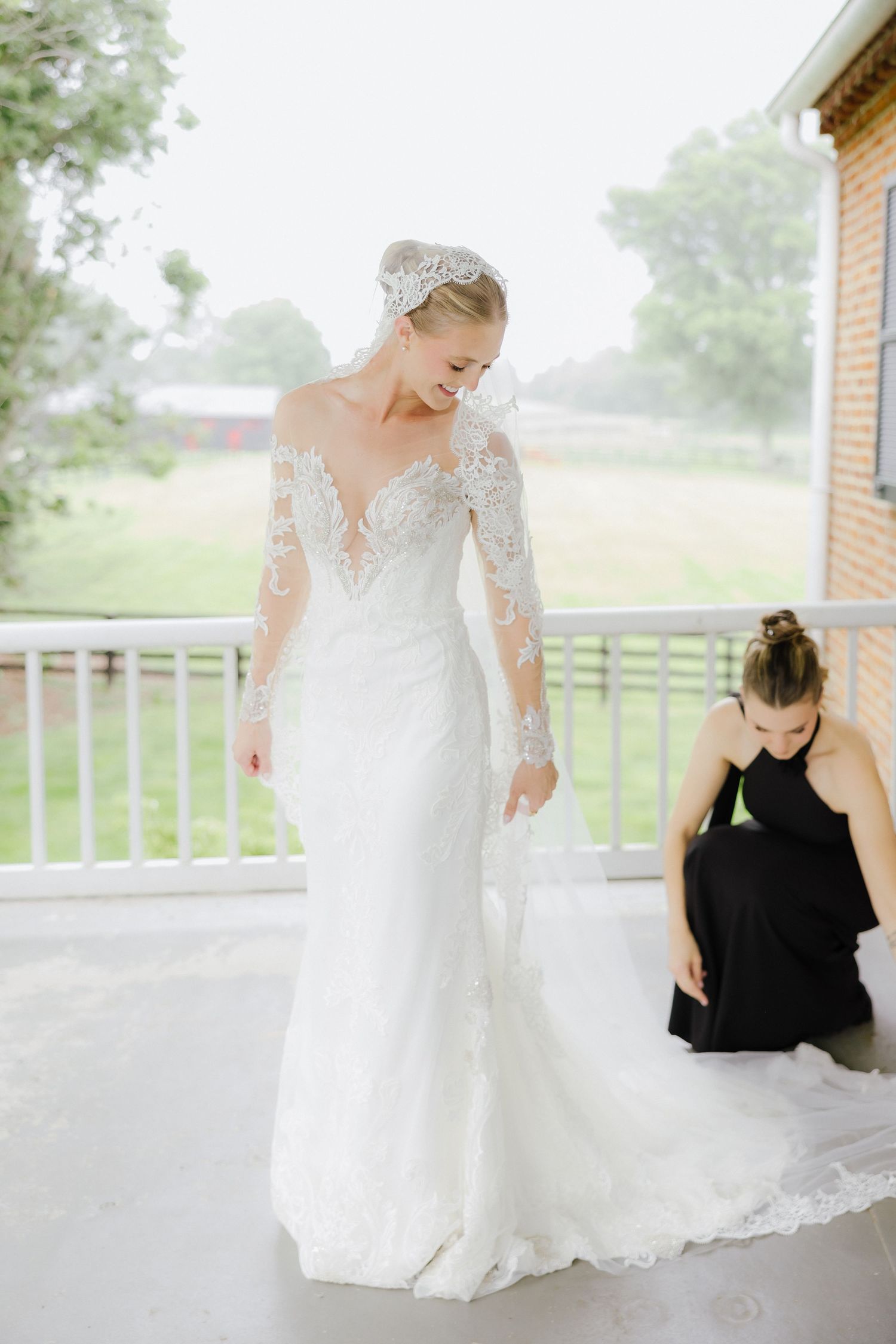 Bride in lace long-sleeve wedding gown being helped by bridesmaid in black dress on outdoor porch.