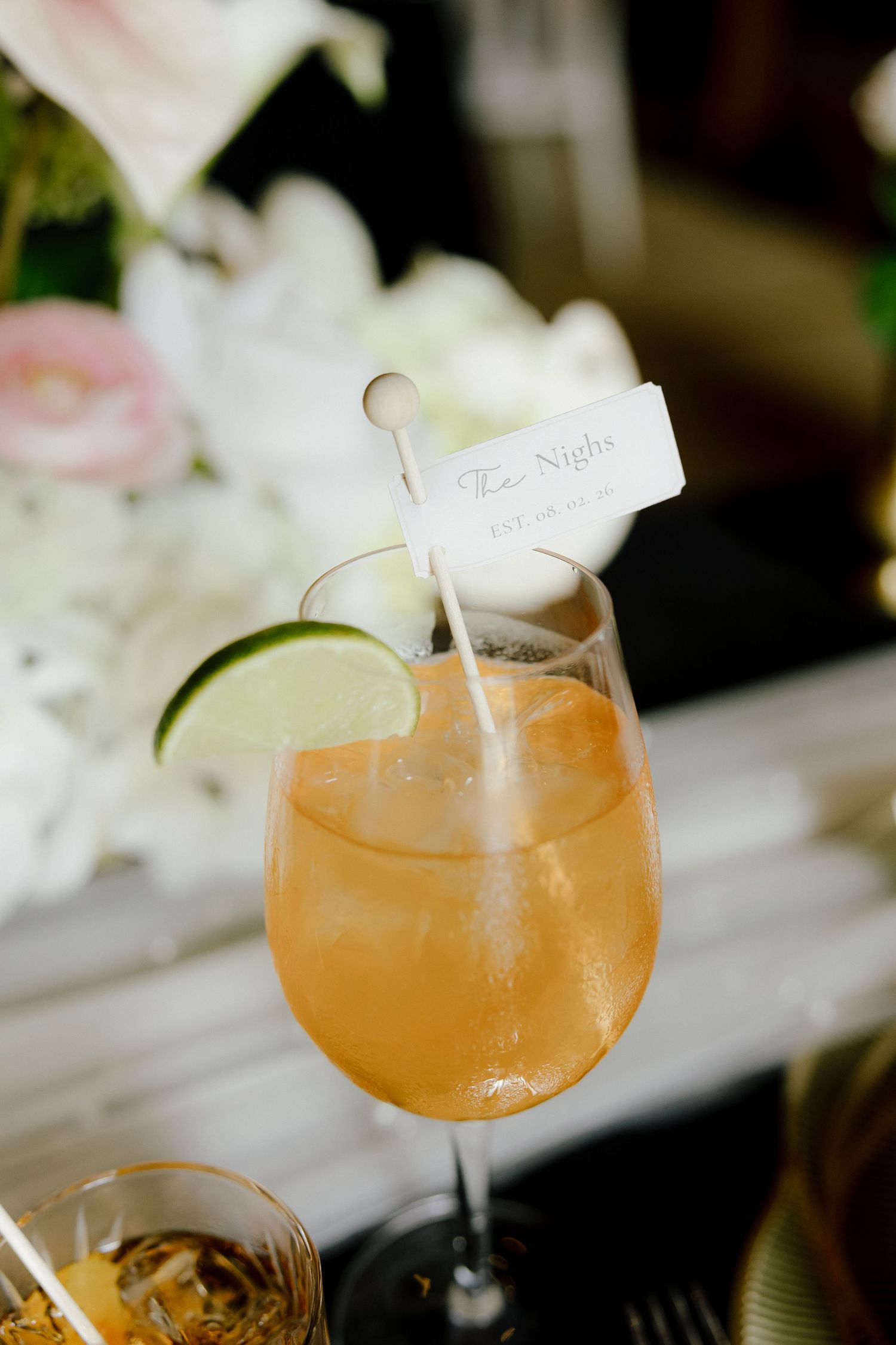 Signature wedding cocktail in wine glass with lime garnish and personalized flag stirrer.