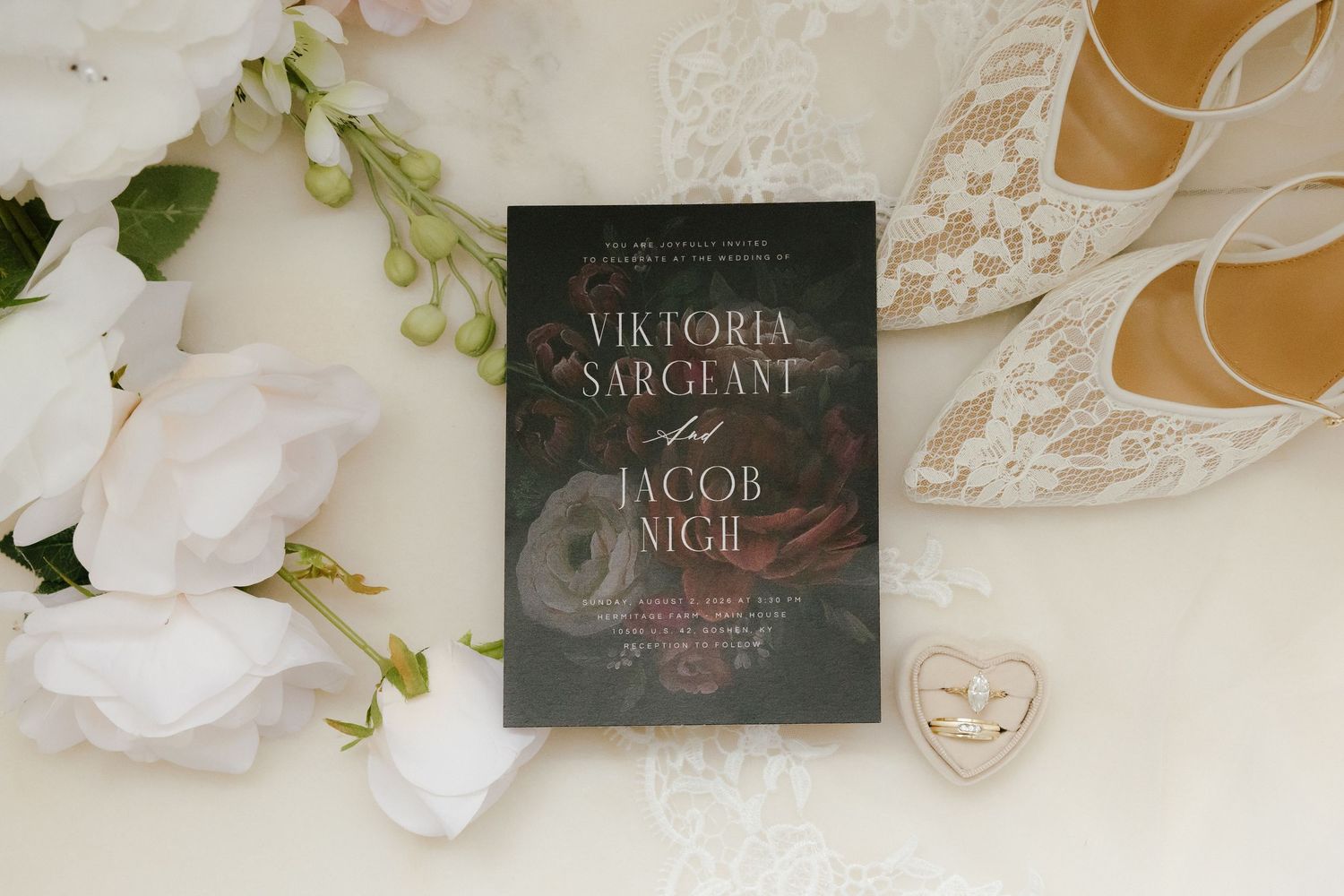Wedding invitation for Viktoria Sargeant and Jacob Nigh displayed with white roses, lace heels, and a heart-shaped ring dish.