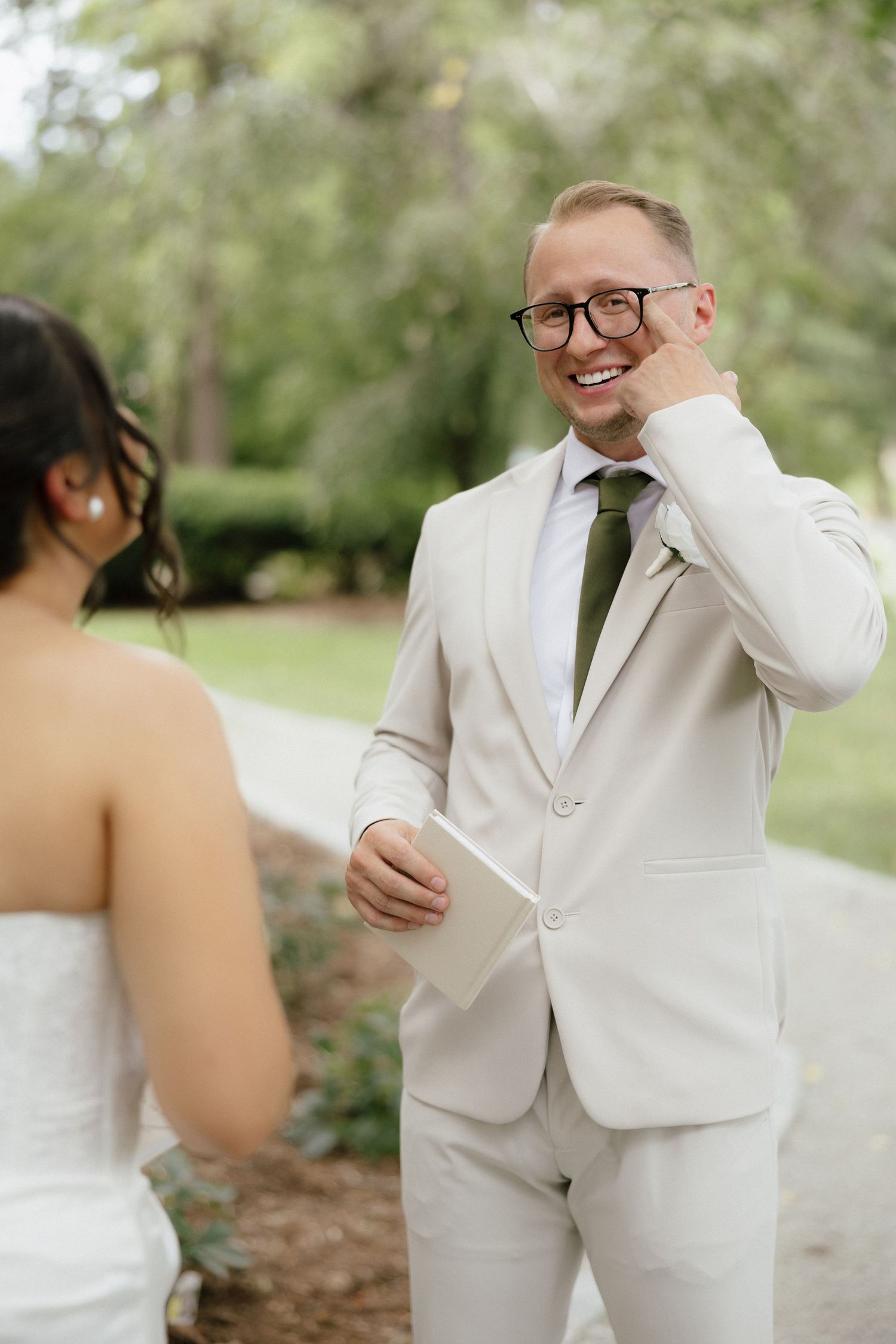 Emotional groom in beige suit with olive tie wiping tears during first look with bride outdoors.