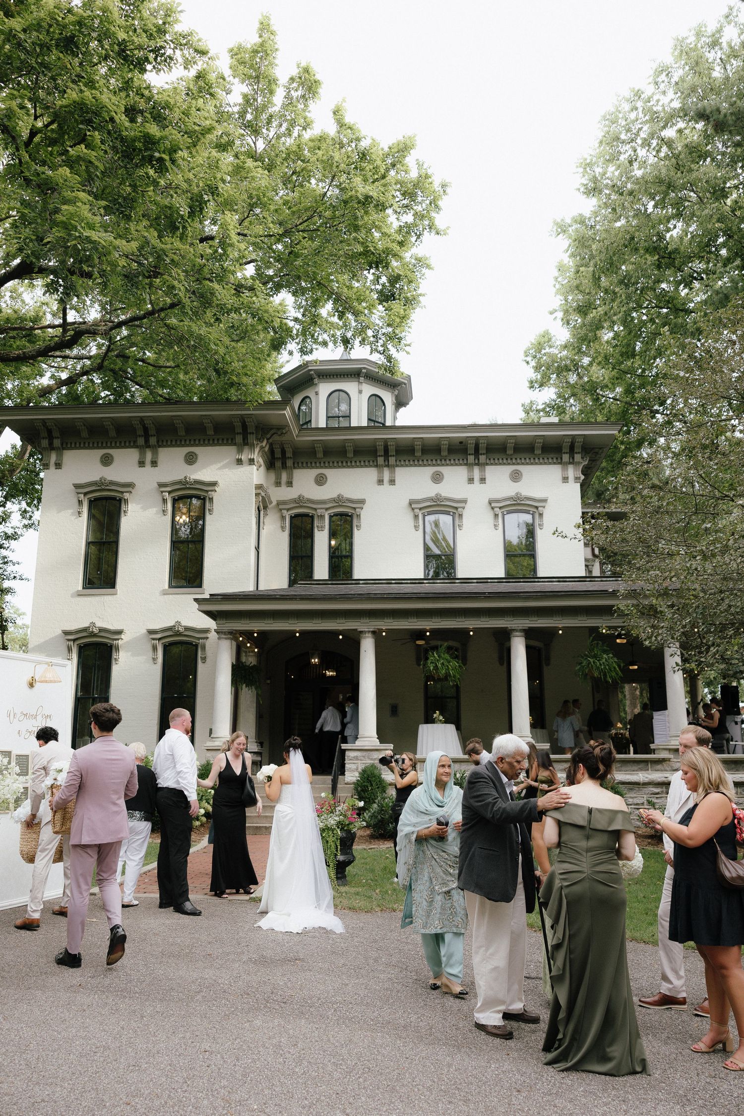 Wedding guests gathered outside a historic white Victorian mansion surrounded by lush green trees.