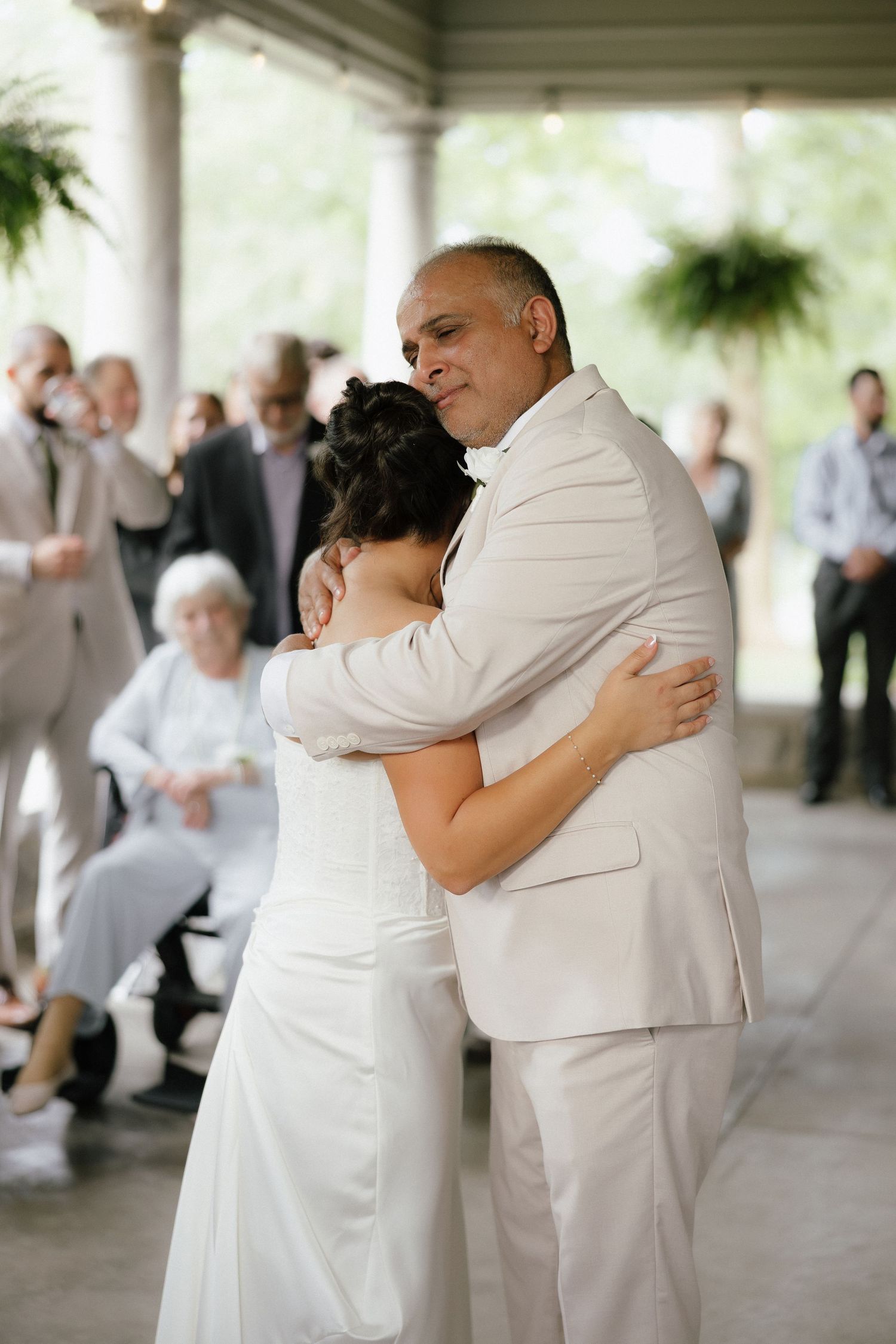 Bride in white gown shares emotional dance with father in cream suit at outdoor wedding reception.