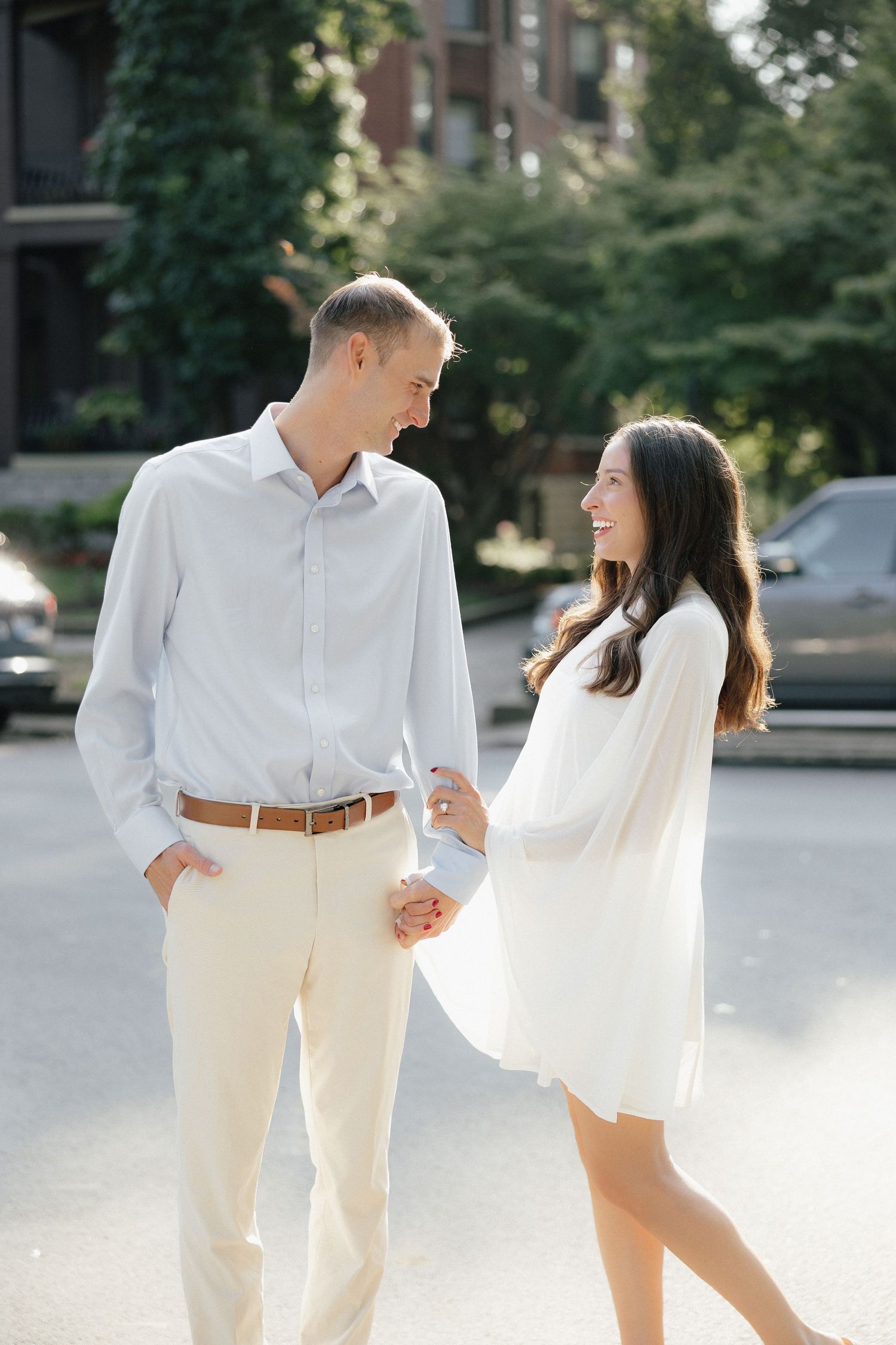 Couple in white outfits holding hands and smiling at each other while walking on a sunny urban street.