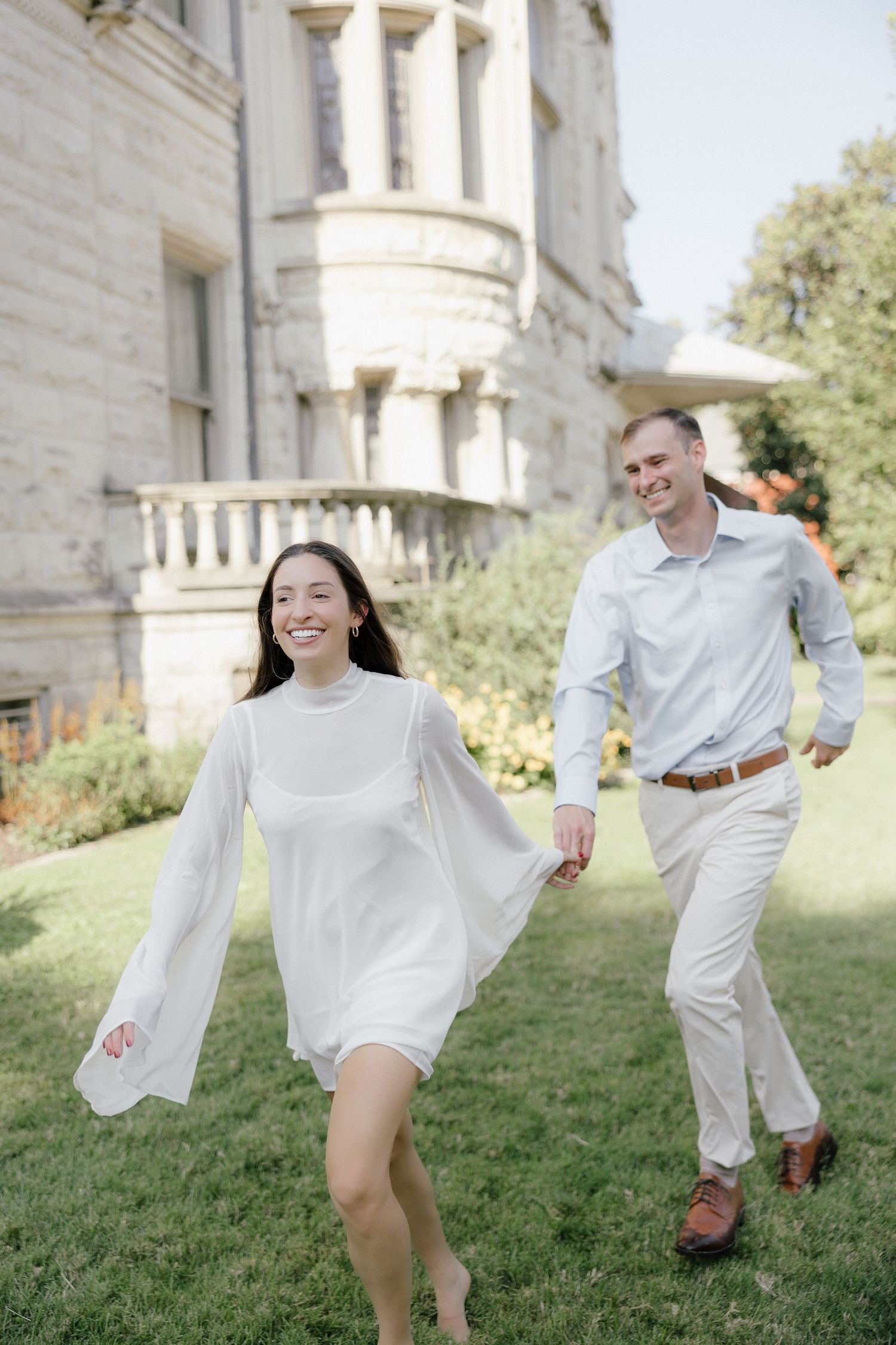 Happy couple holding hands and running on grass in front of a grand historic building on sunny day.