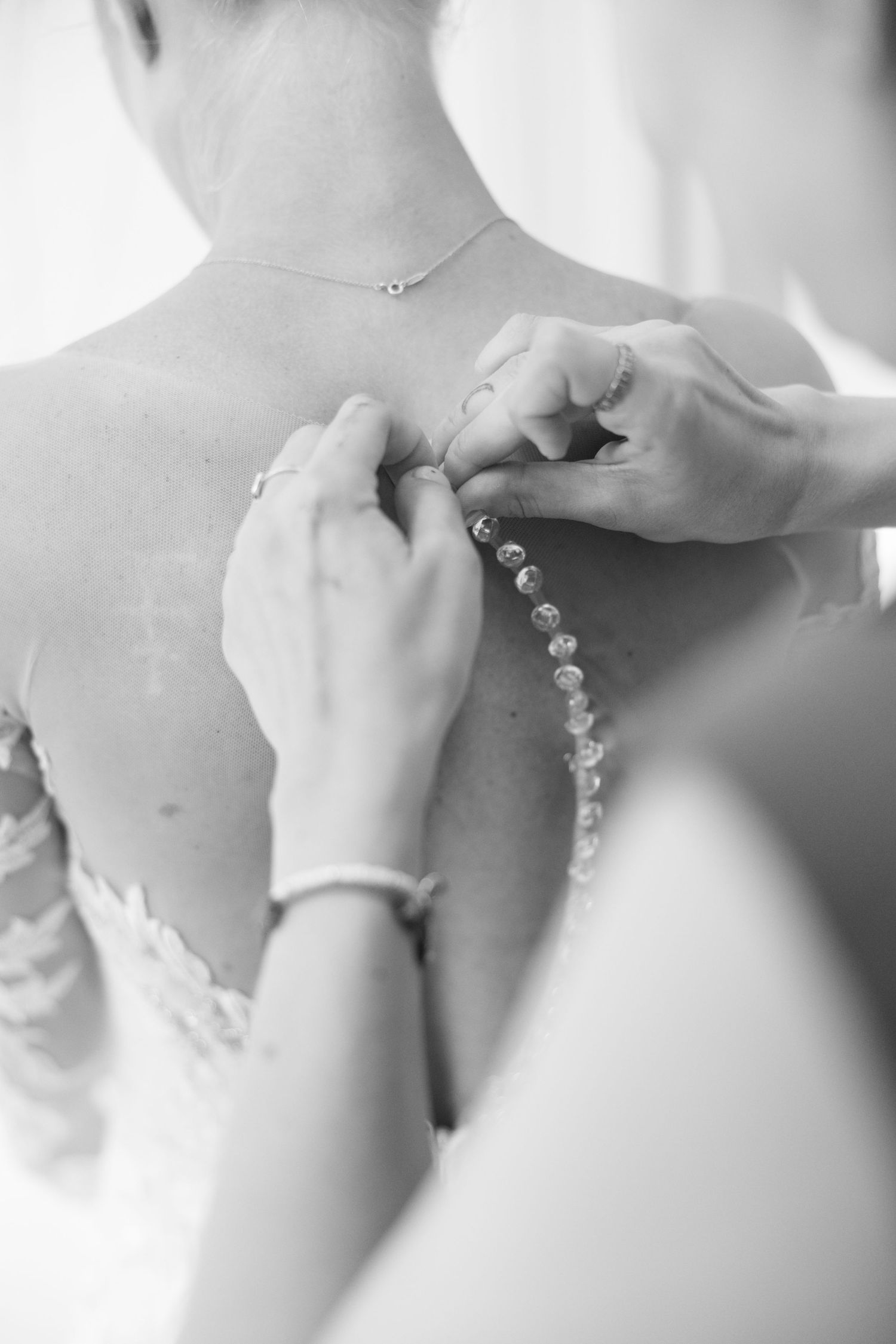 Black and white photo of bridesmaid buttoning pearl buttons on back of lace wedding dress.