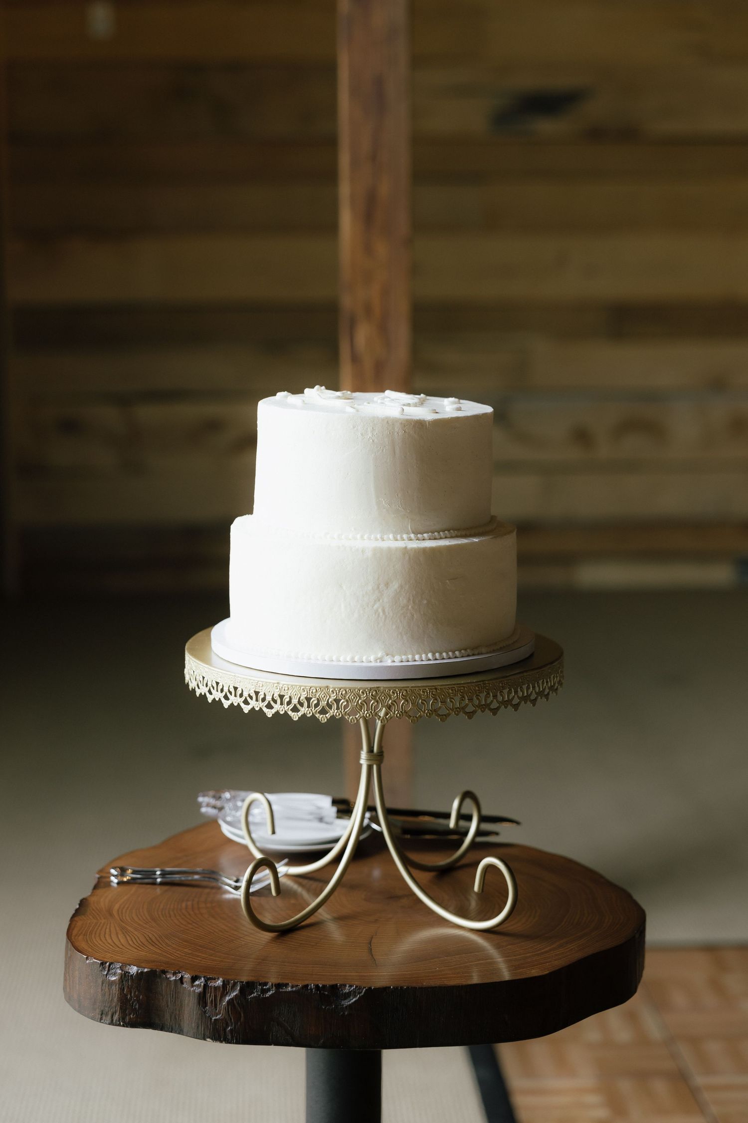 Simple two-tier white wedding cake on gold stand atop wooden slice table in rustic barn venue.