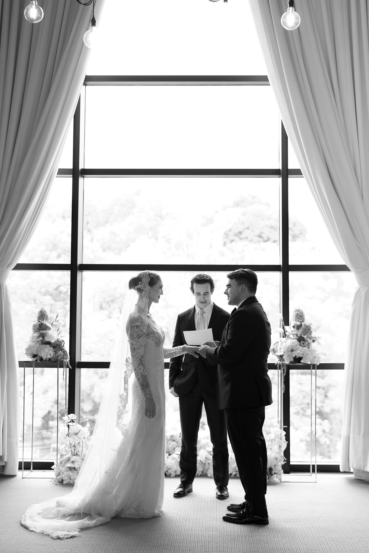 Black and white photo of bride and groom exchanging vows before officiant at elegant wedding ceremony.