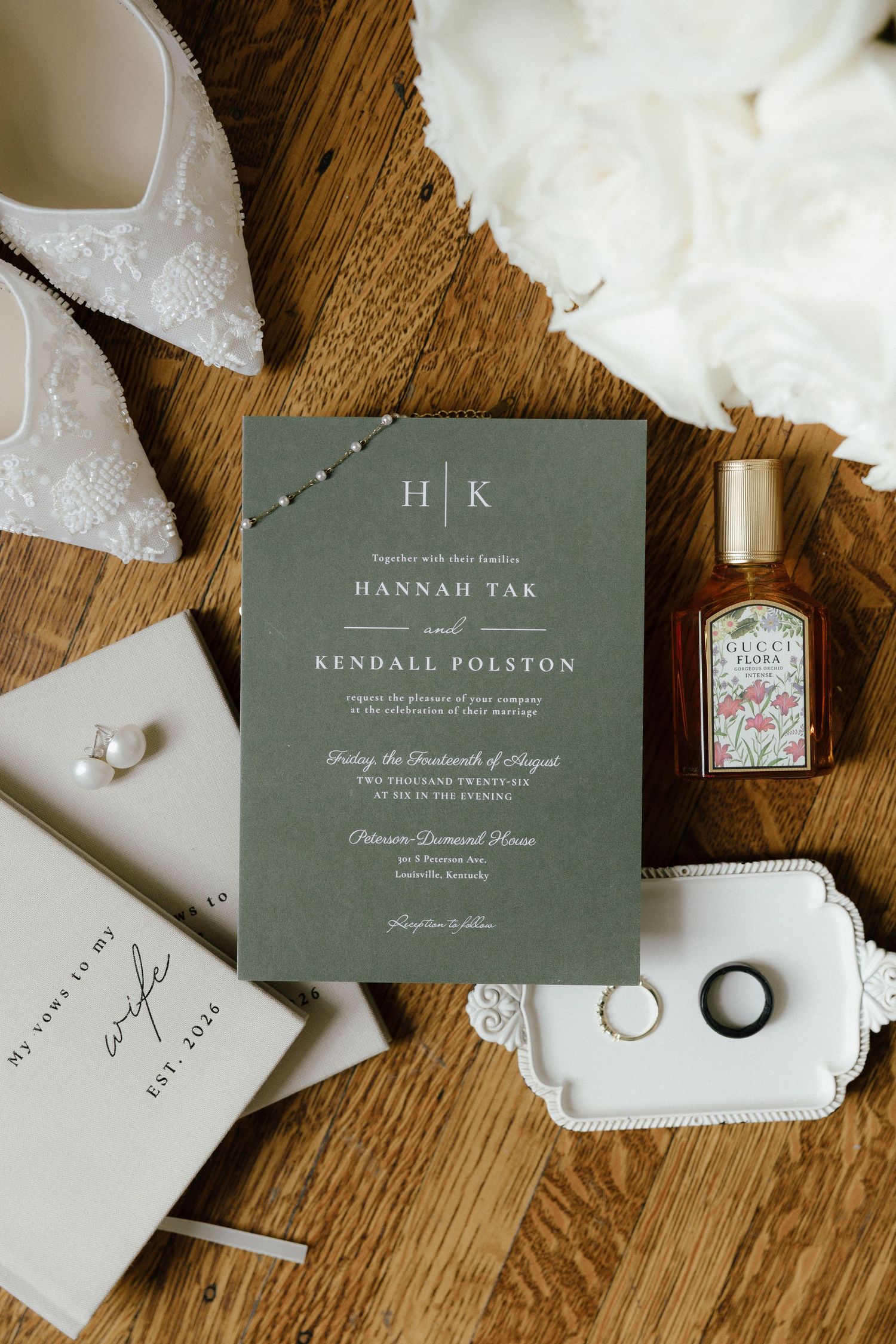 Elegant wedding flat lay with sage green invitation, Gucci Flora perfume, rings, pearl earrings, and lace bridal heels on wood.